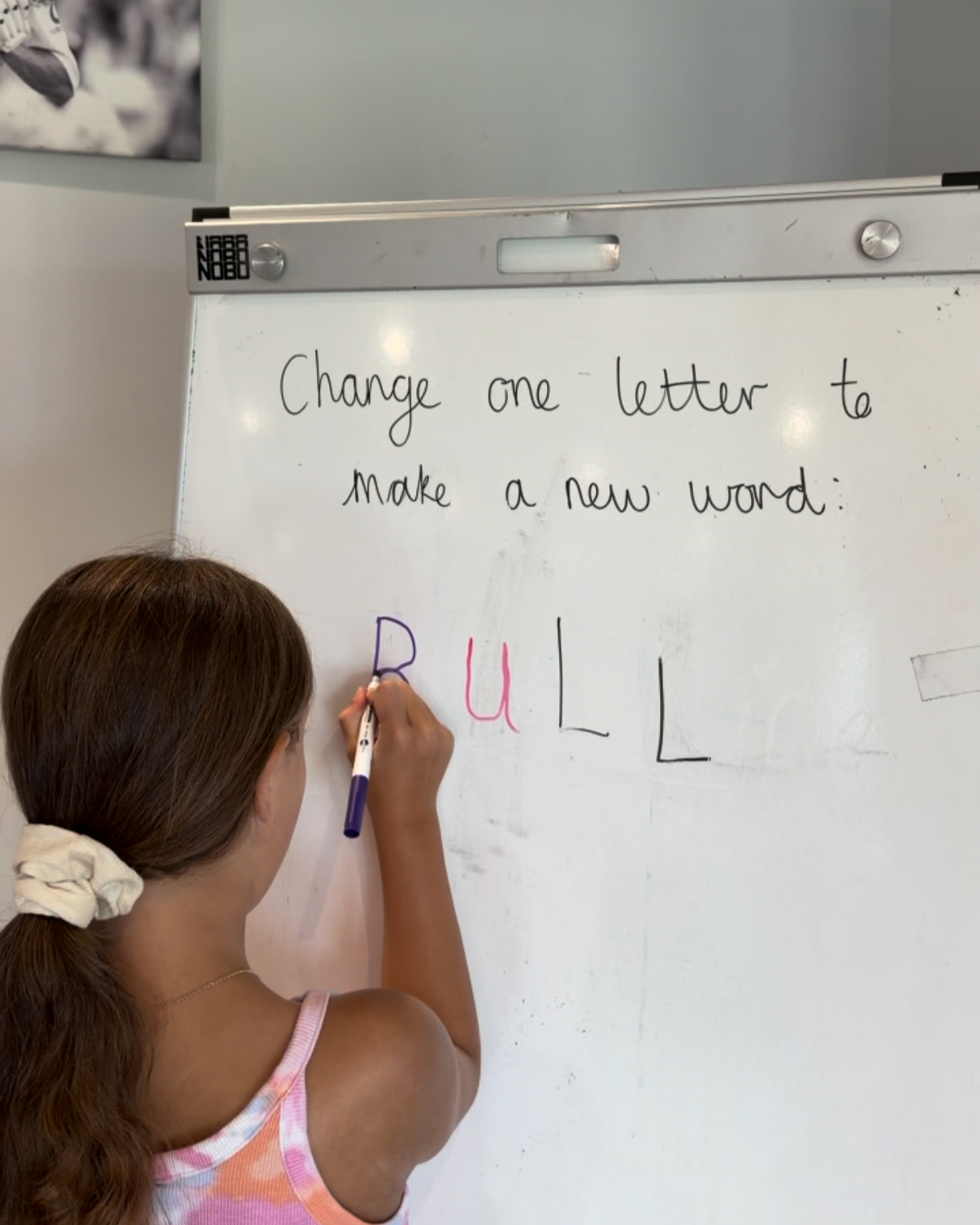A girl with brown hair in a ponytail writing on a whiteboard with a purple marker, changing one letter in the word 'FULL' to make a new word, in a classroom setting.