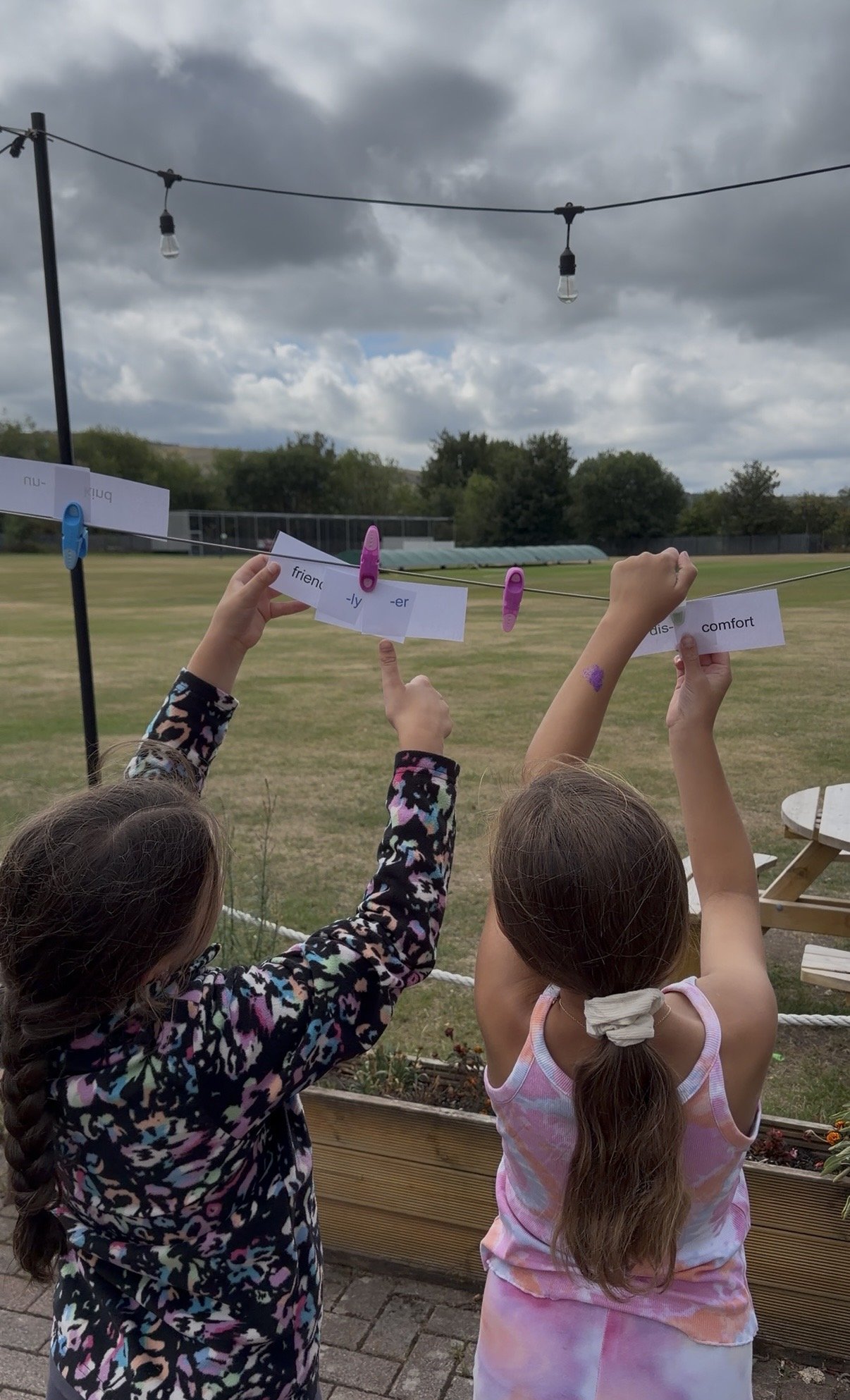 Children hanging paper signs on a clothesline outdoors, with cloudy sky and trees in the background.