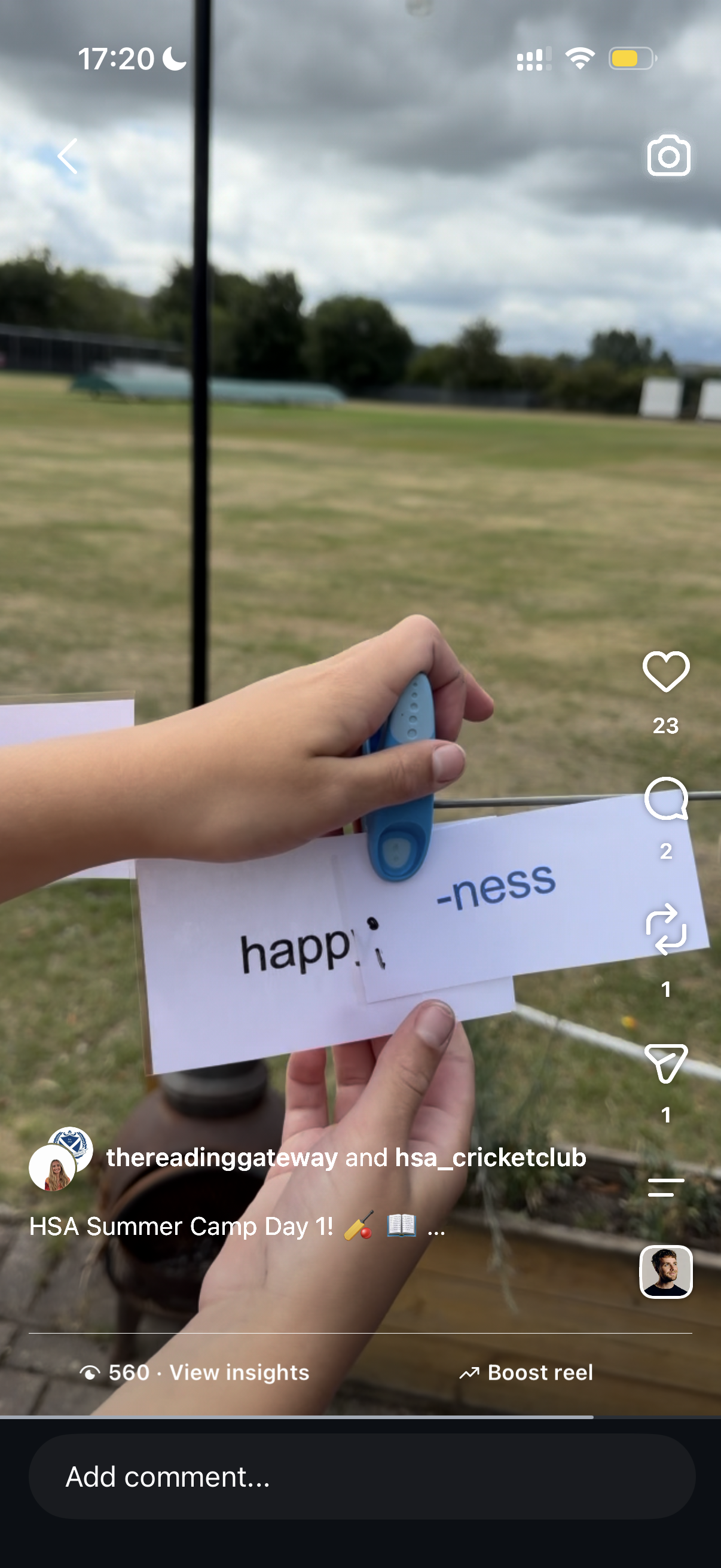 Close-up of a person's hand holding a paper sign with the word 'happiness' missing the letter 'a,' in front of an outdoor scene with grass and trees, under a cloudy sky.