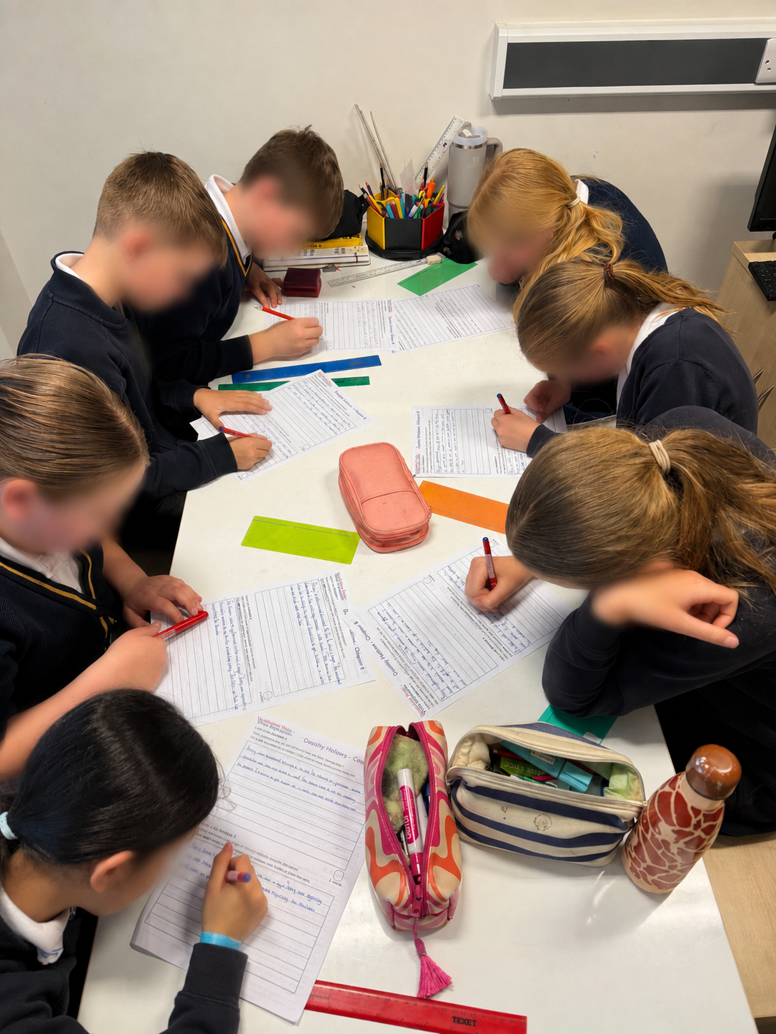 Group of students sitting around a table, working on written assignments with pencils. The table has school supplies including water bottles, pencil cases, and rulers.