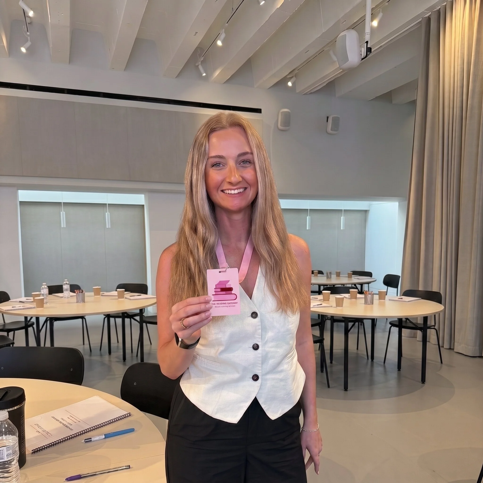 A woman with long blonde hair wearing a sleeveless white top and black pants holding a pink booklet and standing in a conference room. The room has round tables with notebooks, pens, water bottles, and coffee cups, with large windows and beige curtains in the background.