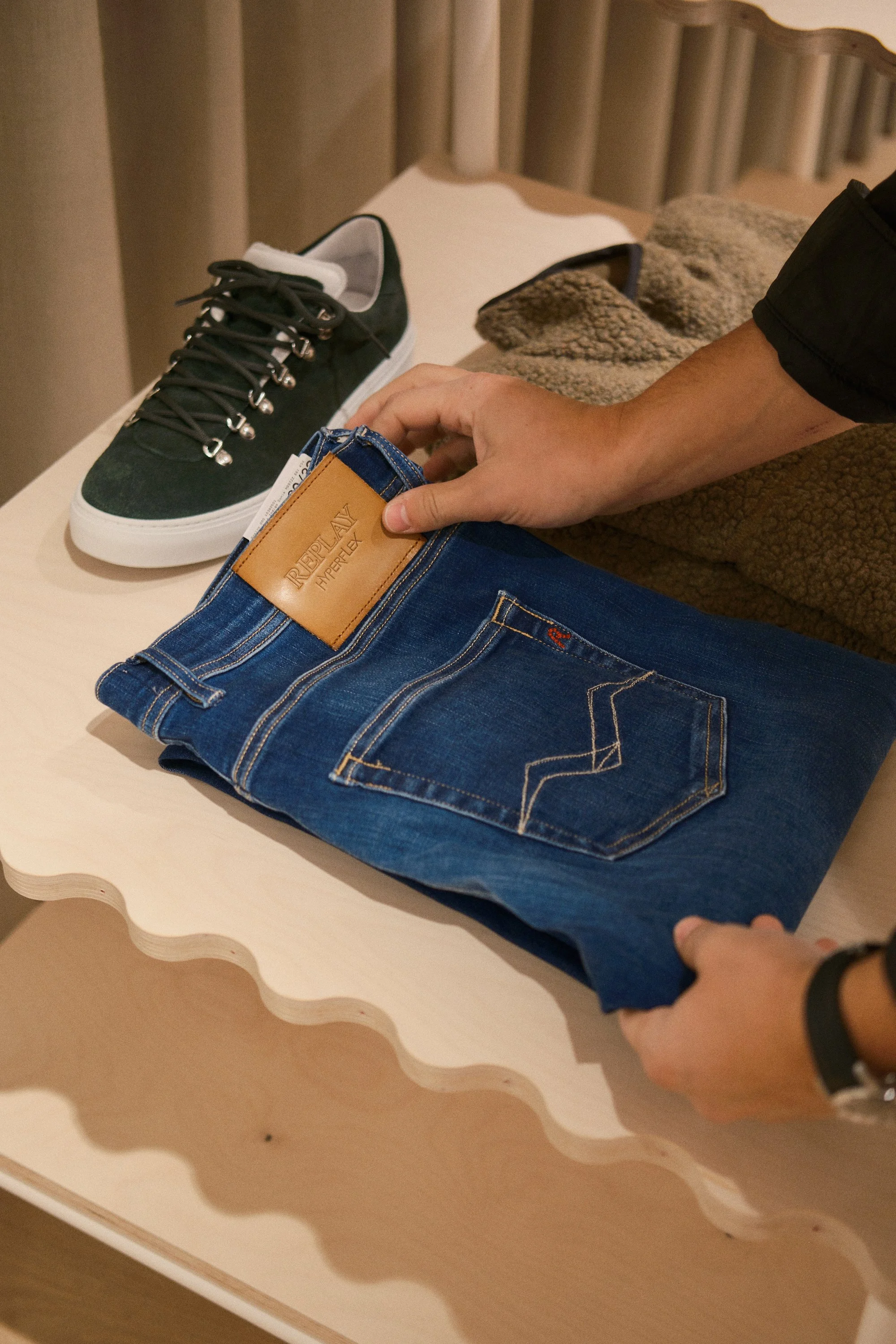 Et par sko og et belte ligger på et bord. Handen til en person holder på et par jeans med merket "Replay".