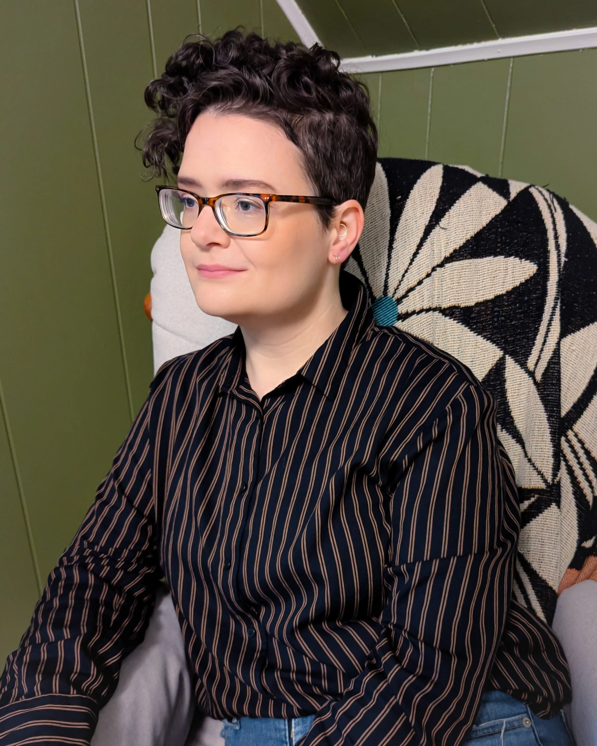 A smiling nonbinary person with short curly hair and glasses sitting in a chair. | Virtual therapy for ADHD in Massachusetts