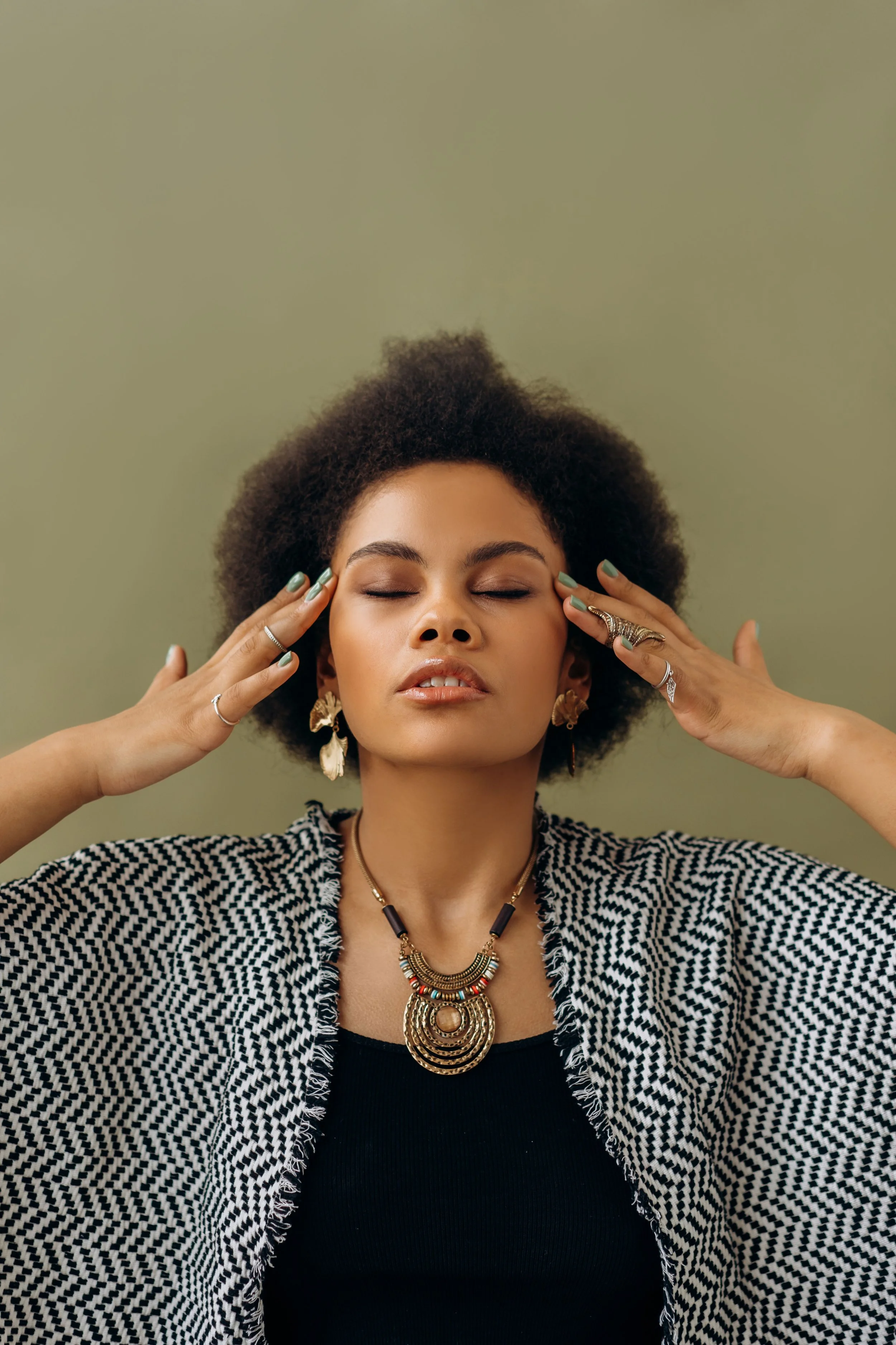 A feminine-presenting person of color with their eyes closed and their fingertips at their temples, as if in meditation