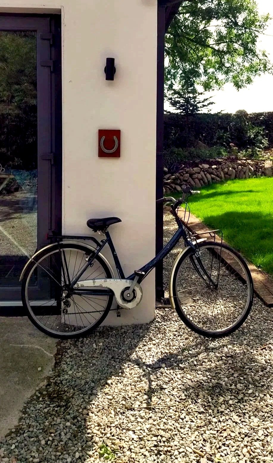 A vintage bicycle with a front basket, white tires, and a black frame, leaning against a white wall outside a building with a glass door. The background features a well-maintained green lawn, trees, and a stone border.