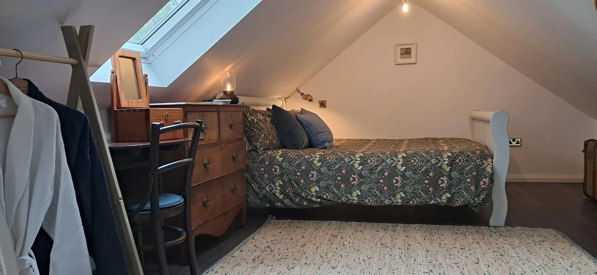 A cozy attic bedroom with a sloped ceiling, featuring a bed with a floral patterned bedspread and four pillows, a wooden dresser, a small mirror, a lamp, a skylight window, and a beige carpet.