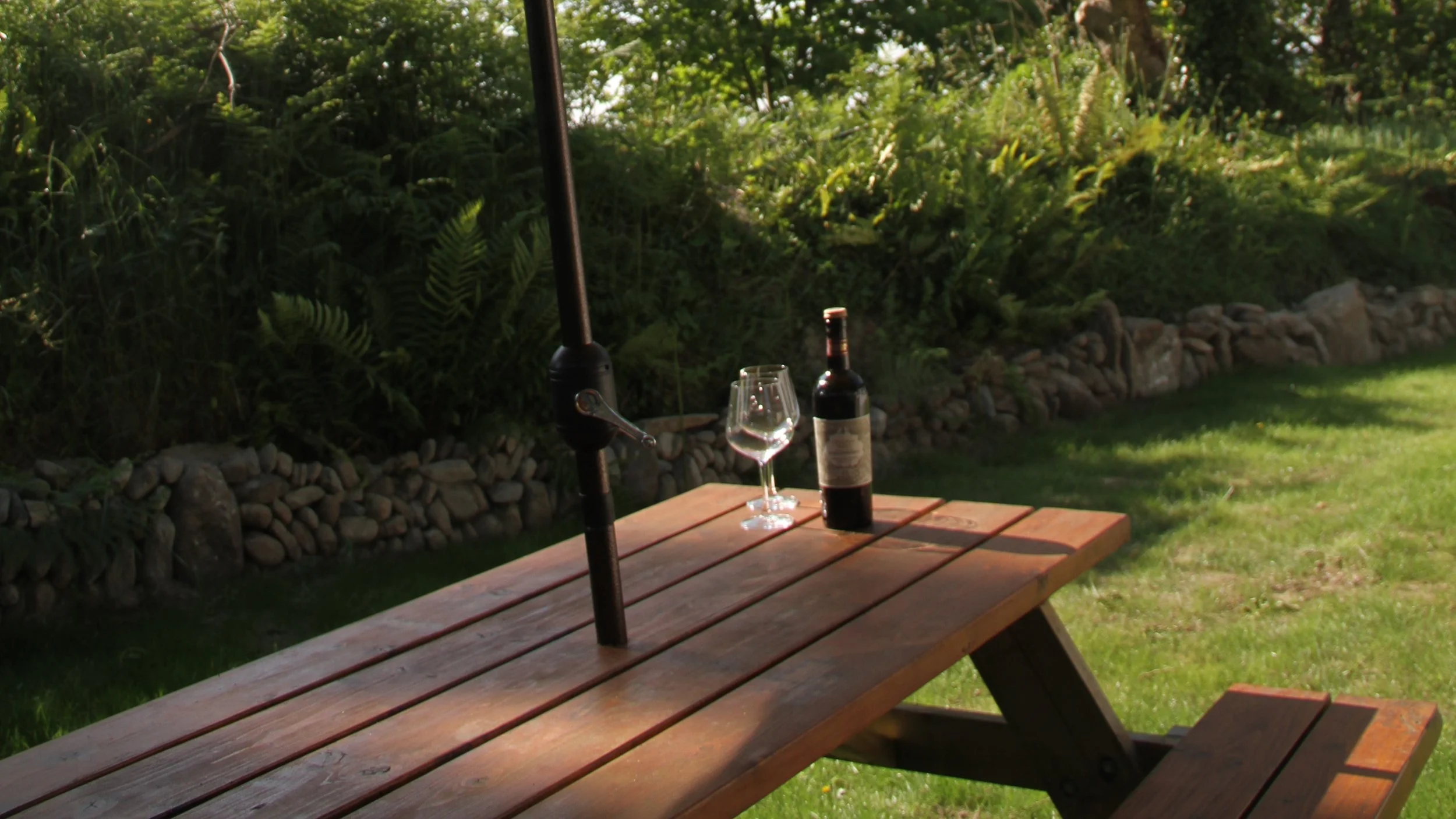 A wooden picnic table with a bottle of red wine, an empty wine glass, and a wine opener on top, set outdoors on green grass with a stone wall and lush green bushes in the background.