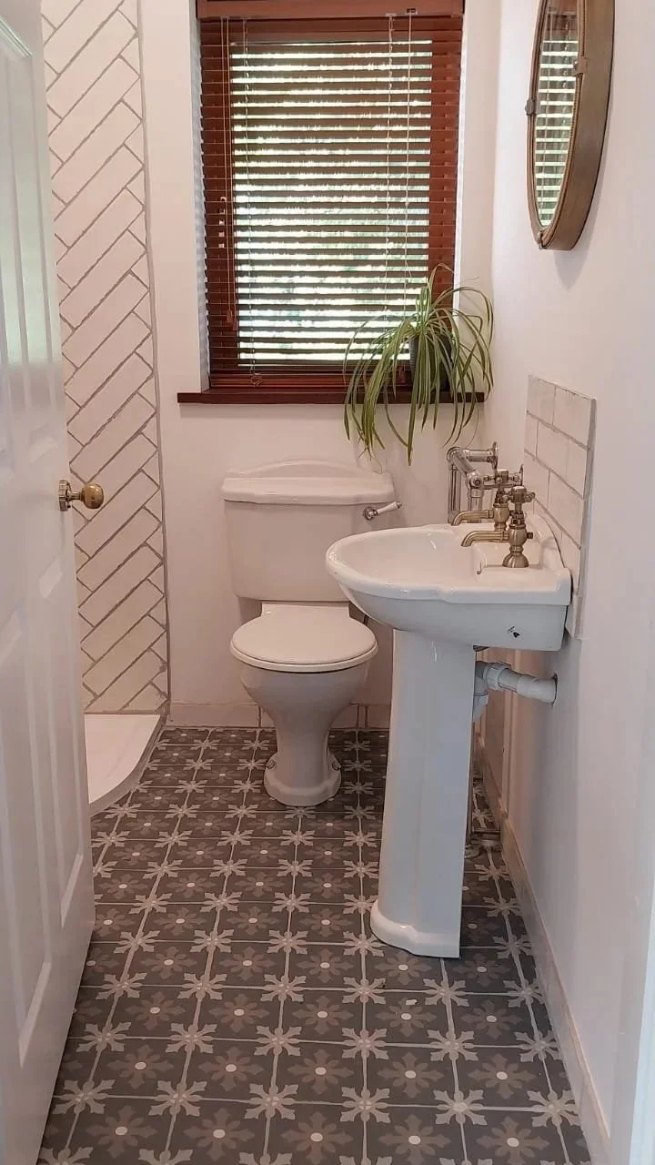 Small bathroom with a toilet and a pedestal sink, a window with wooden blinds, a wall-mounted mirror, a plant on the window sill, and patterned floor tiles.