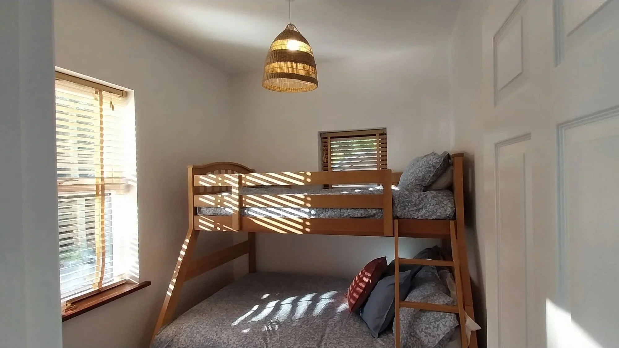 A bedroom with two vertical windows with blinds on white walls, a wooden loft bed with bedding, and sunlight casting shadows through the blinds.