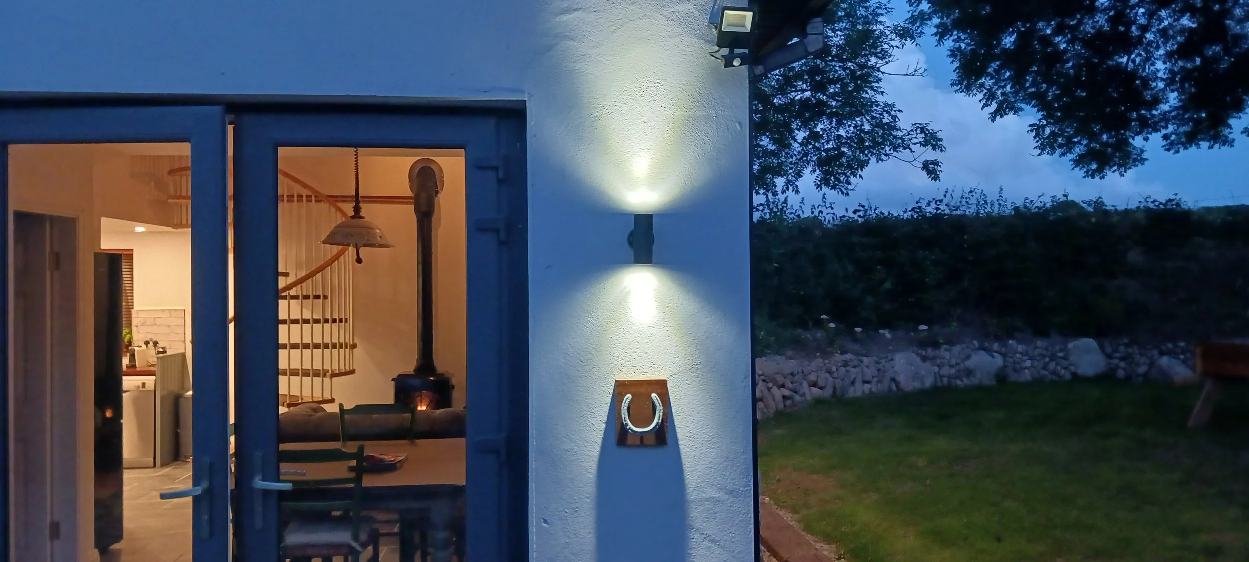 A view of an open glass door leading into a warmly lit home with stairs and a hanging lamp inside. Outside, there is a wall-mounted light, a horseshoe decoration, a grassy area, trees, and a cloudy blue evening sky.