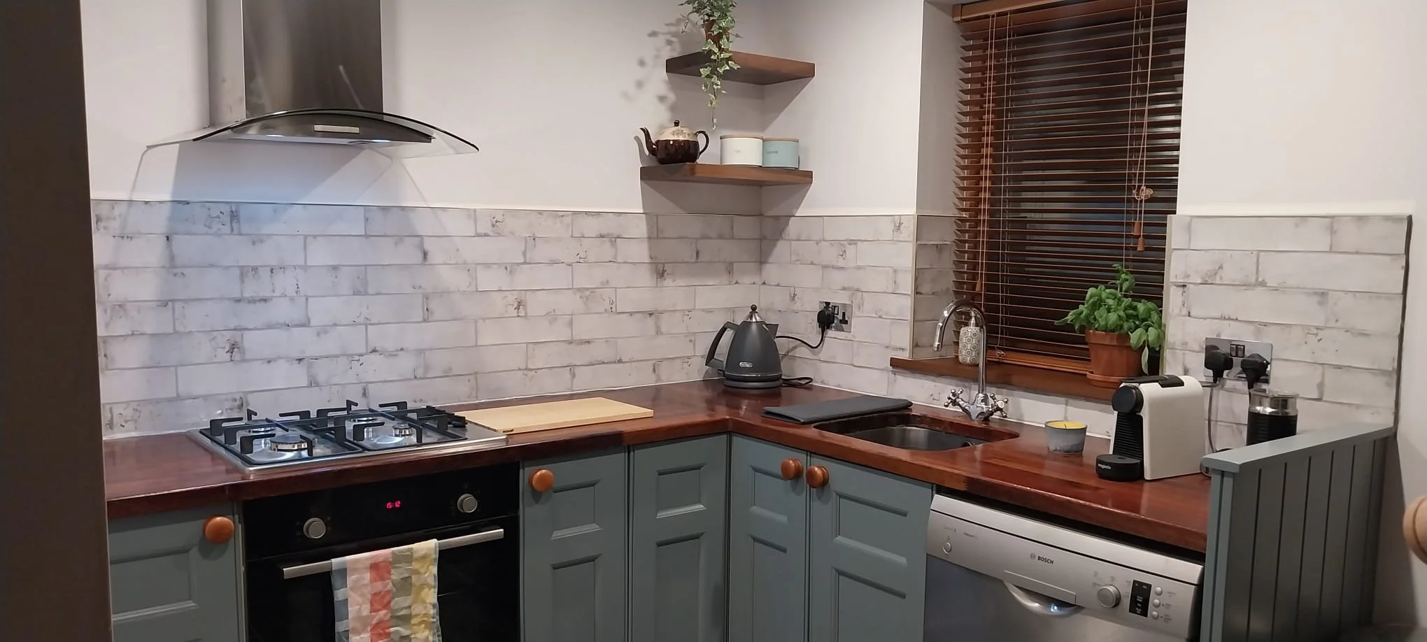 A kitchen with light blue cabinets, a wooden countertop, a gas stove, a kettle, a sink, a potted plant on the window sill, a coffee machine, and a washing machine. There are wooden shelves above the counter and wooden blinds on the window.