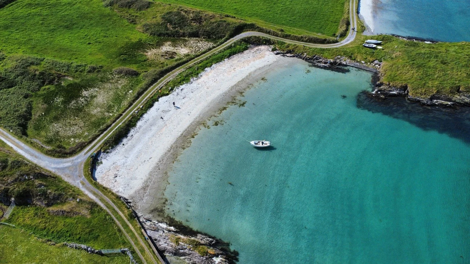 A small boat anchored near a pebble beach in a calm, turquoise cove with green grassy hills surrounding it and a narrow paved road leading to the water. Two cars and a boat are parked on the grassy area near the water.