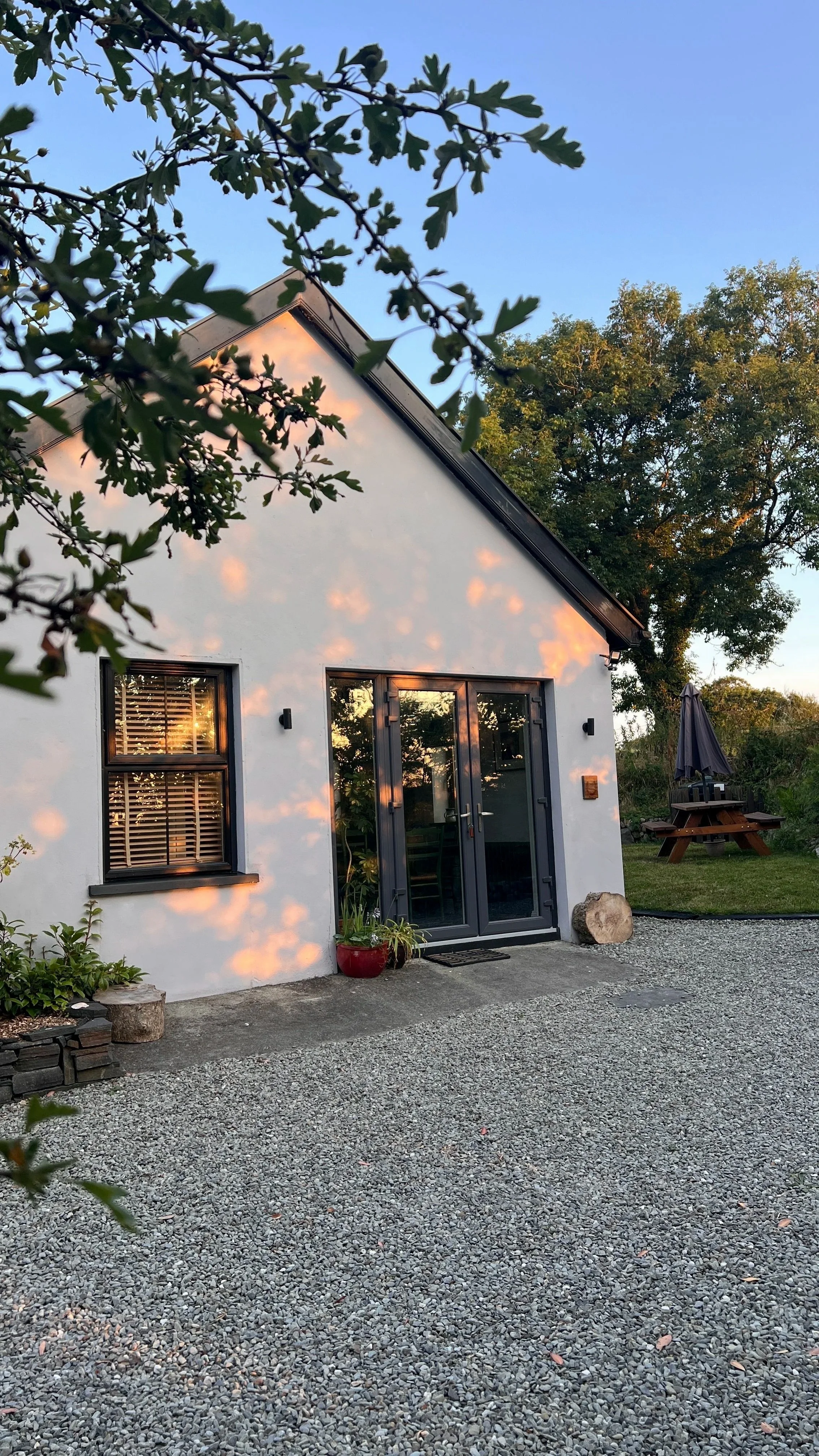 A white house with black window frames and a gey door, surrounded by trees and a gravel yard, with a picnic table with an umbrella in the background.