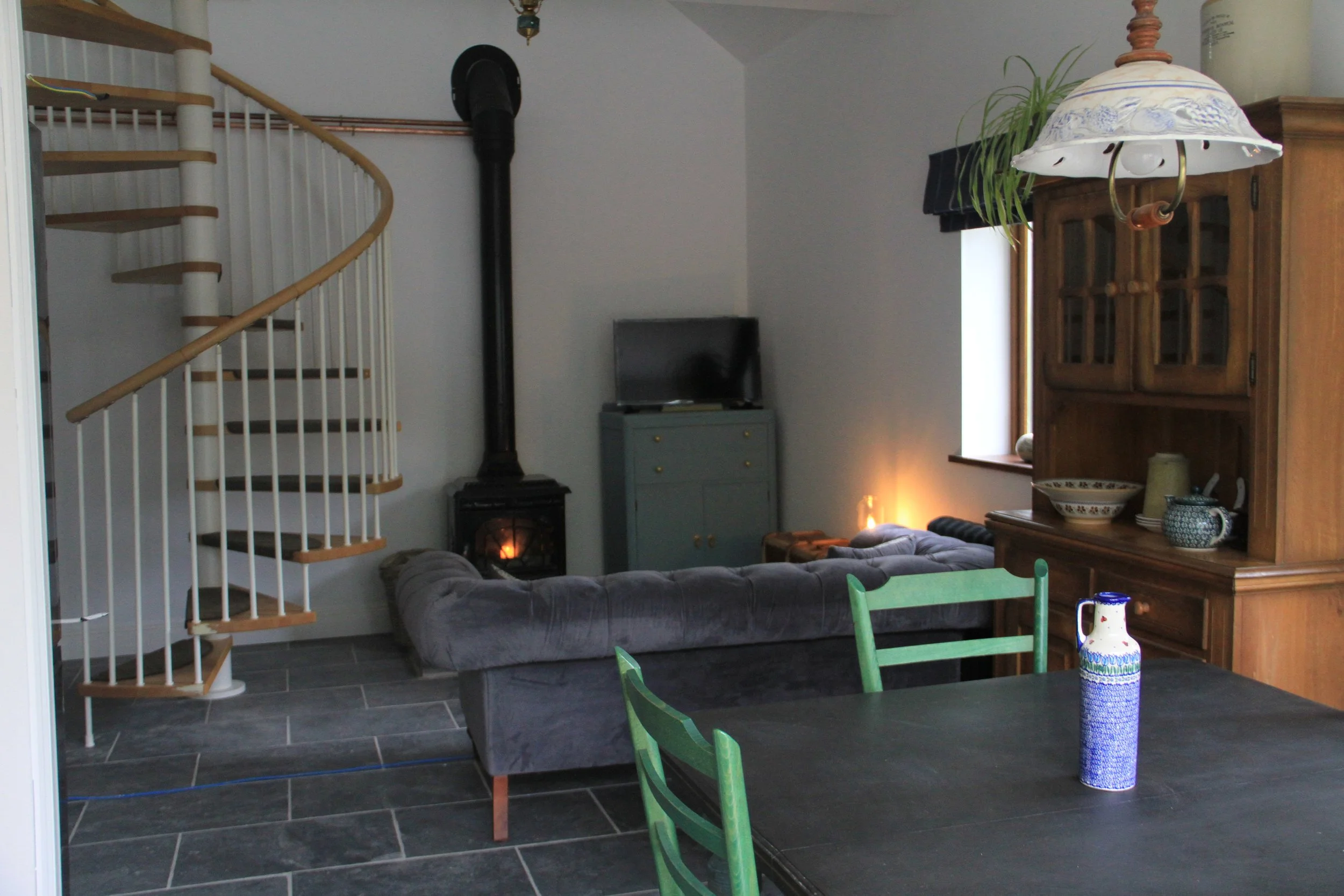 Living room with a spiral staircase on the left, a gray couch, a small black stove with a lit flame, a small gray cabinet with a TV on top, a wooden cabinet with glass doors, a window with black curtains, a hanging lamp with floral patterns, a green 