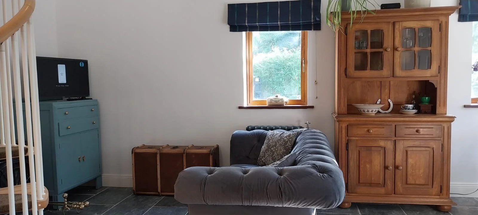 A cozy living room with a gray tufted sofa, wooden cabinet, blue chest of drawers, and a window with a dark valance.