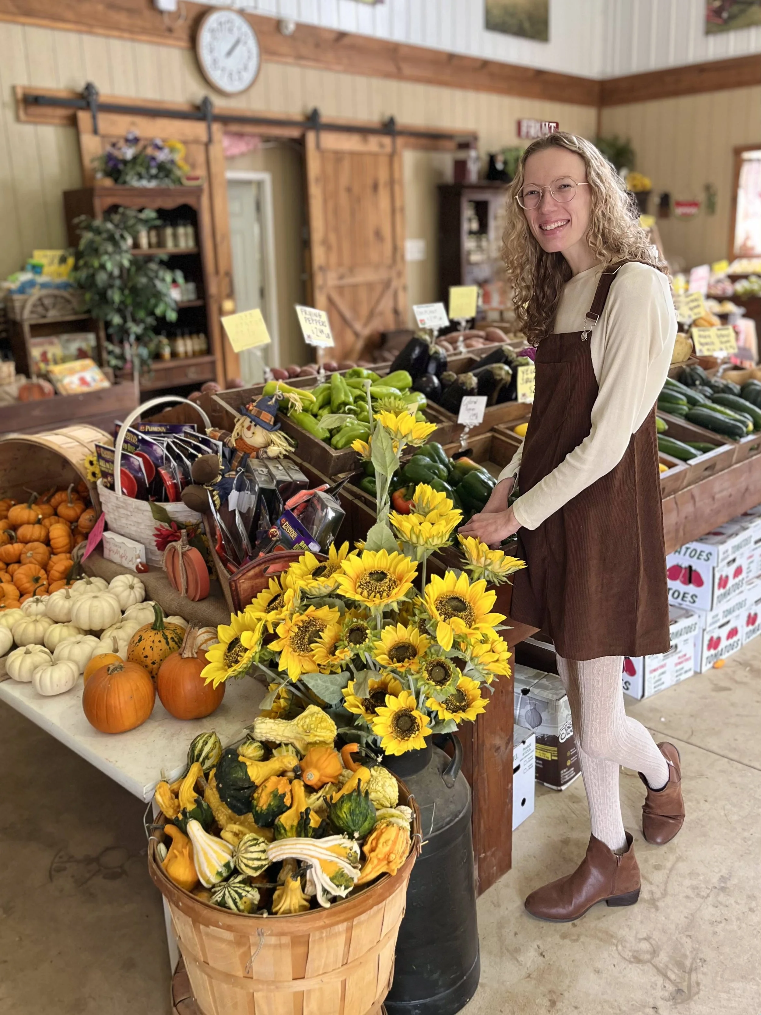 A woman with curly blond hair, glasses, wearing a cream long-sleeve top, brown apron, white lace tights, and brown boots, smiling while shopping for vegetables at a grocery store with pumpkins, gourds, sunflower arrangements, and other produce on dis
