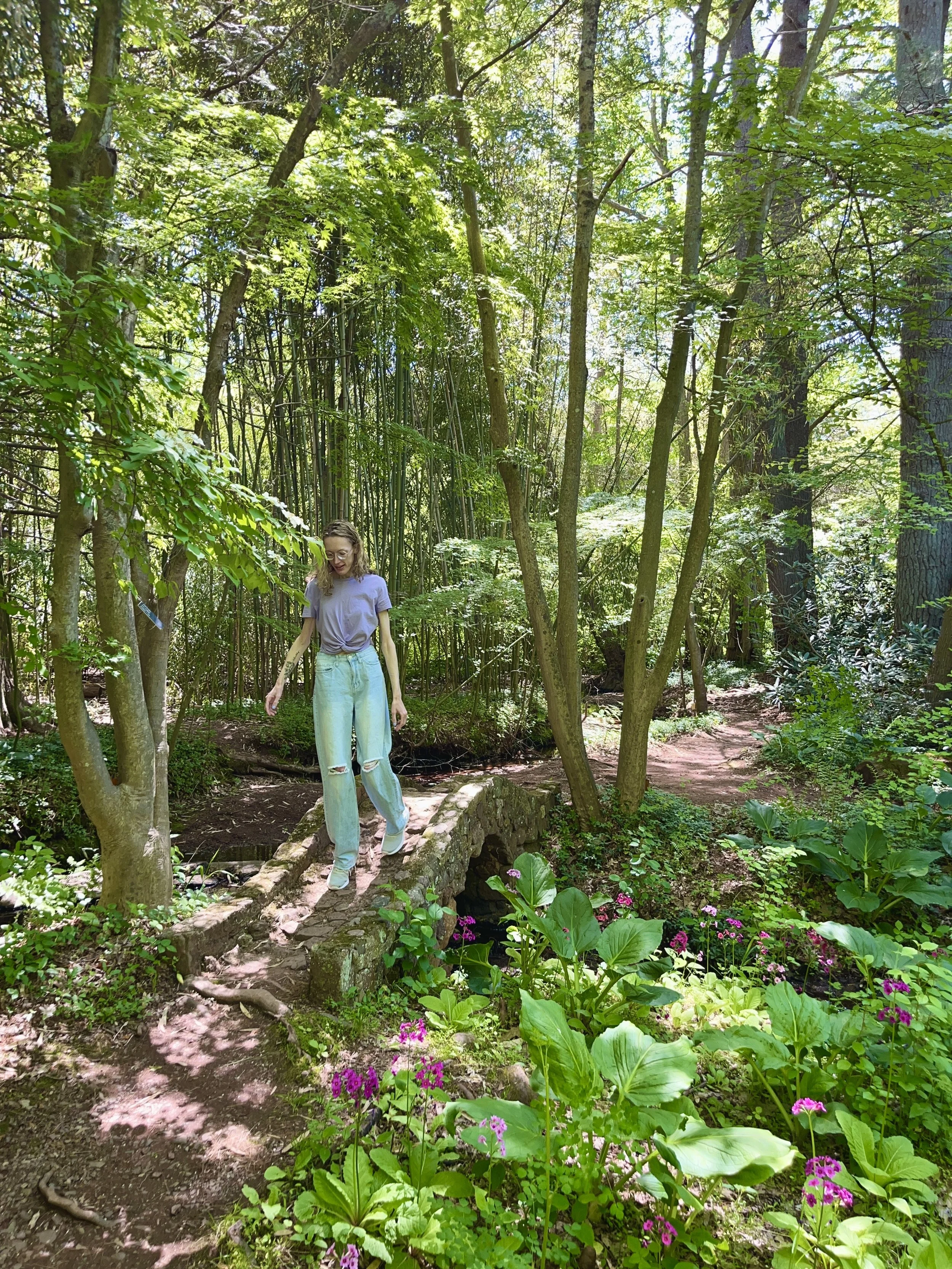 A woman walking on a small stone footbridge through a lush green forest filled with trees and flowering plants.