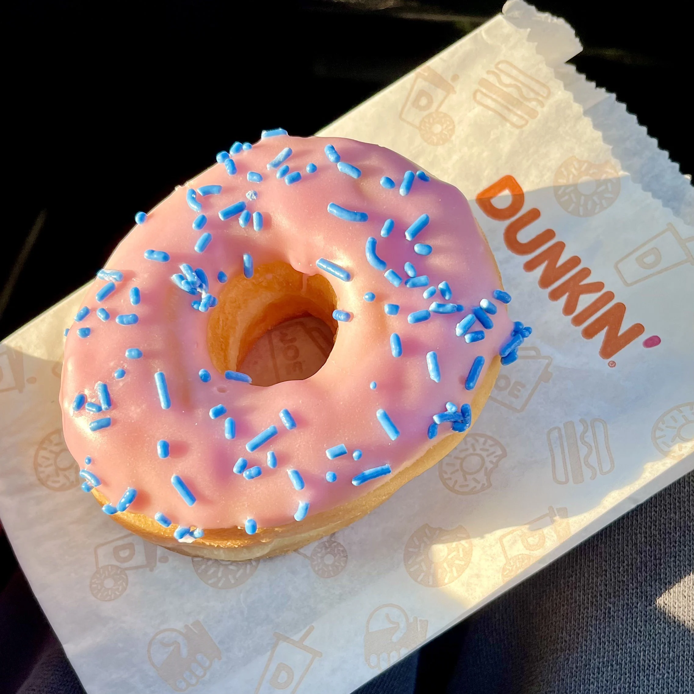 Close-up of a pink donut with blue sprinkles on top with a partially visible orange and white packaging in the background.