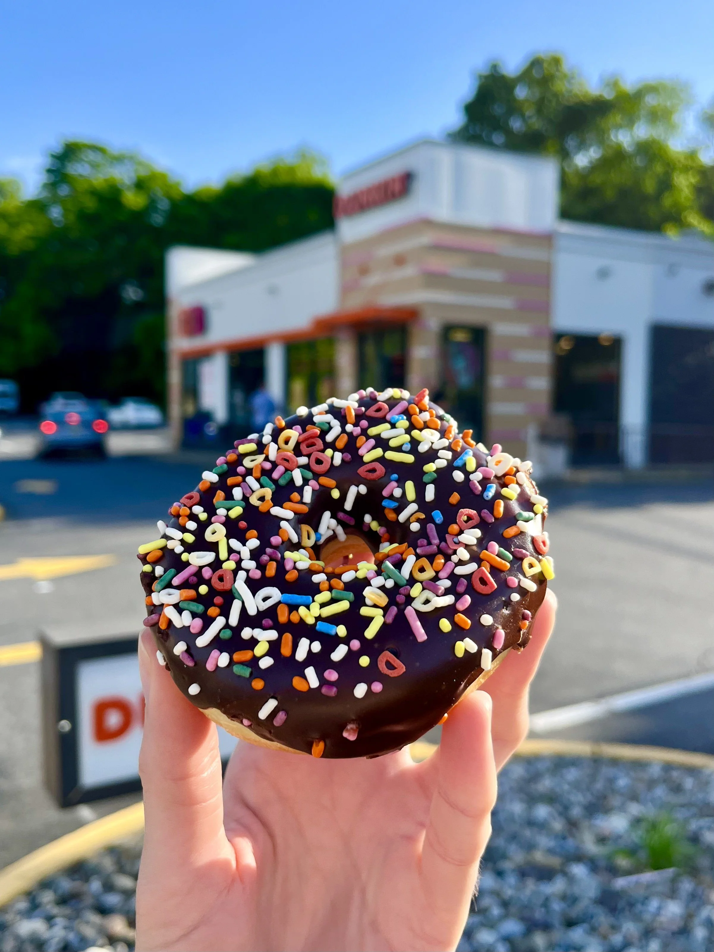 A person holding a chocolate donut with colorful sprinkles in front of a fast food restaurant with white and pink exterior, set against a bright blue sky and green trees.