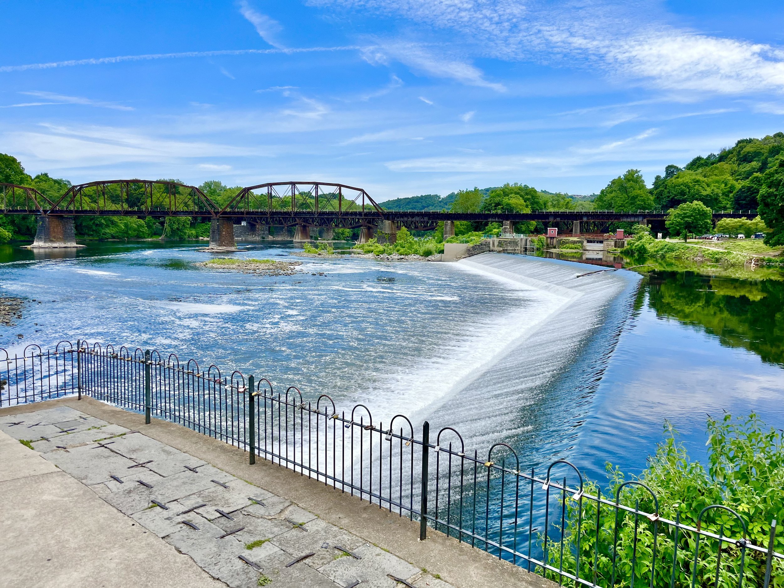 A metal bridge over a river surrounded by green trees under a blue sky with white clouds.