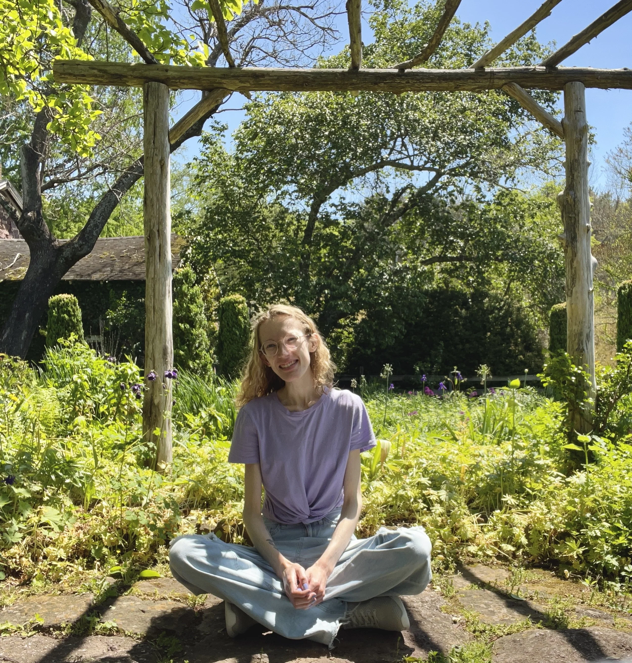 A woman with glasses and curly hair sitting cross-legged on a stone path in a garden with green plants, trees, and purple flowers, smiling at the camera, outdoors on a sunny day.