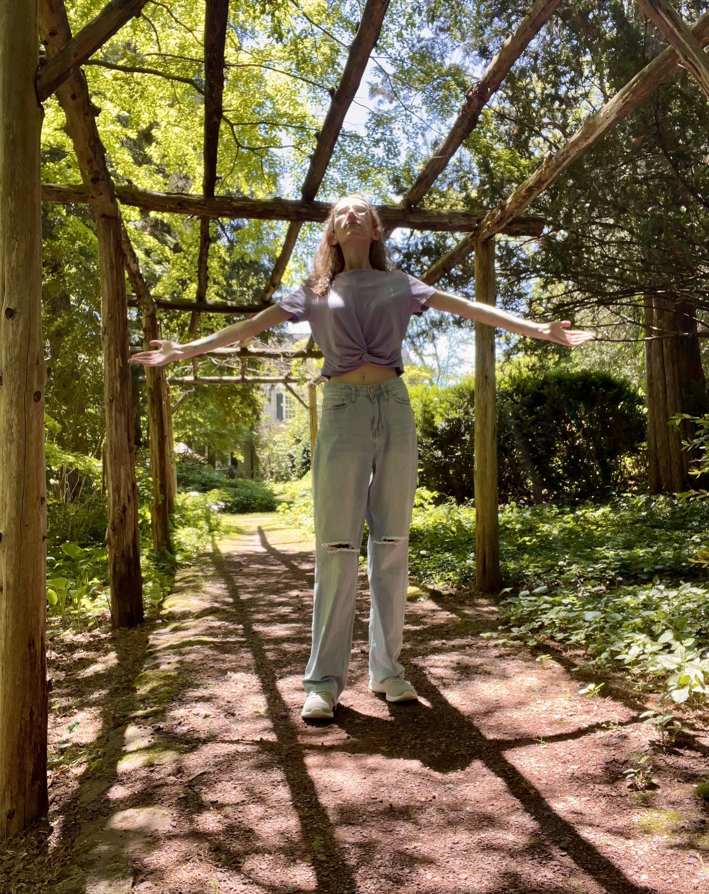 A young woman standing outdoors on a dirt pathway amidst a leafy green environment, with her arms open wide and her head tilted back, under a wooden structure made of logs. Sunlight filters through the trees, creating shadows on the ground.