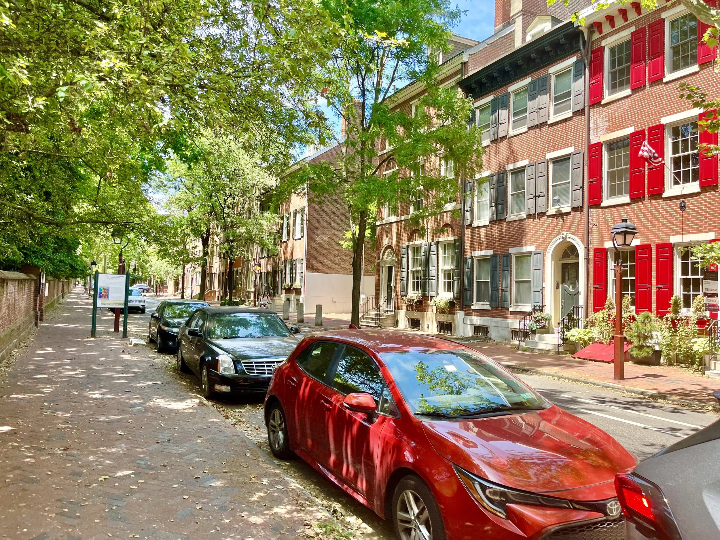 Street view with parked cars, trees with green leaves, and red brick buildings with black and red shutters, and a blue sky.