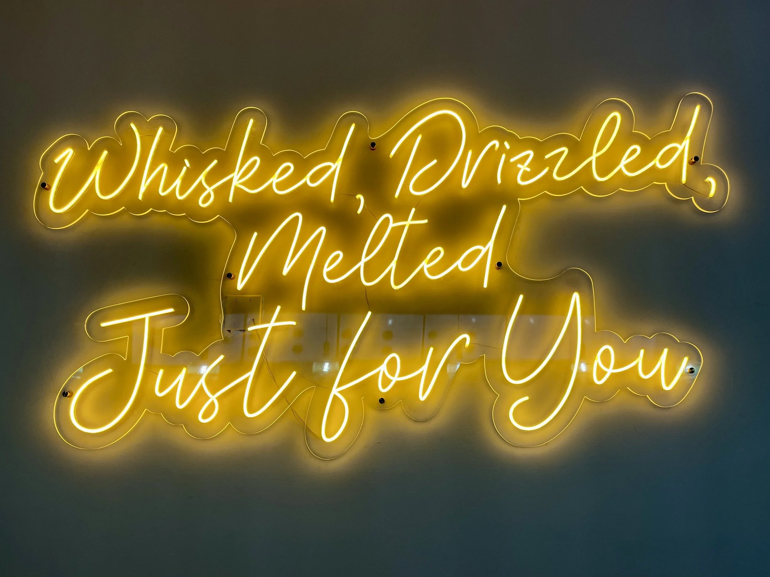 Neon sign reading 'Whisked, Drizzled, Melted, Just for You' in yellow cursive light lettering.