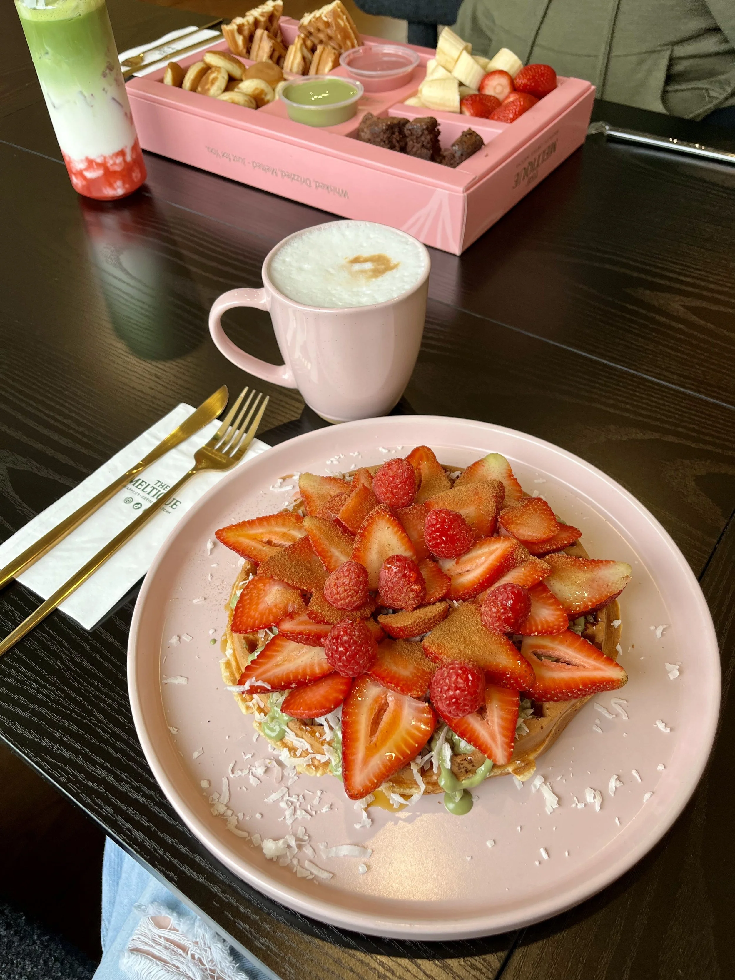 A plate of strawberry shortcake topped with slices of strawberries and raspberries, with shredded coconut around the edges. A pink mug of latte with foam and a caramel drizzle is nearby. In the background, a pink tray holds various snacks and fruit, 