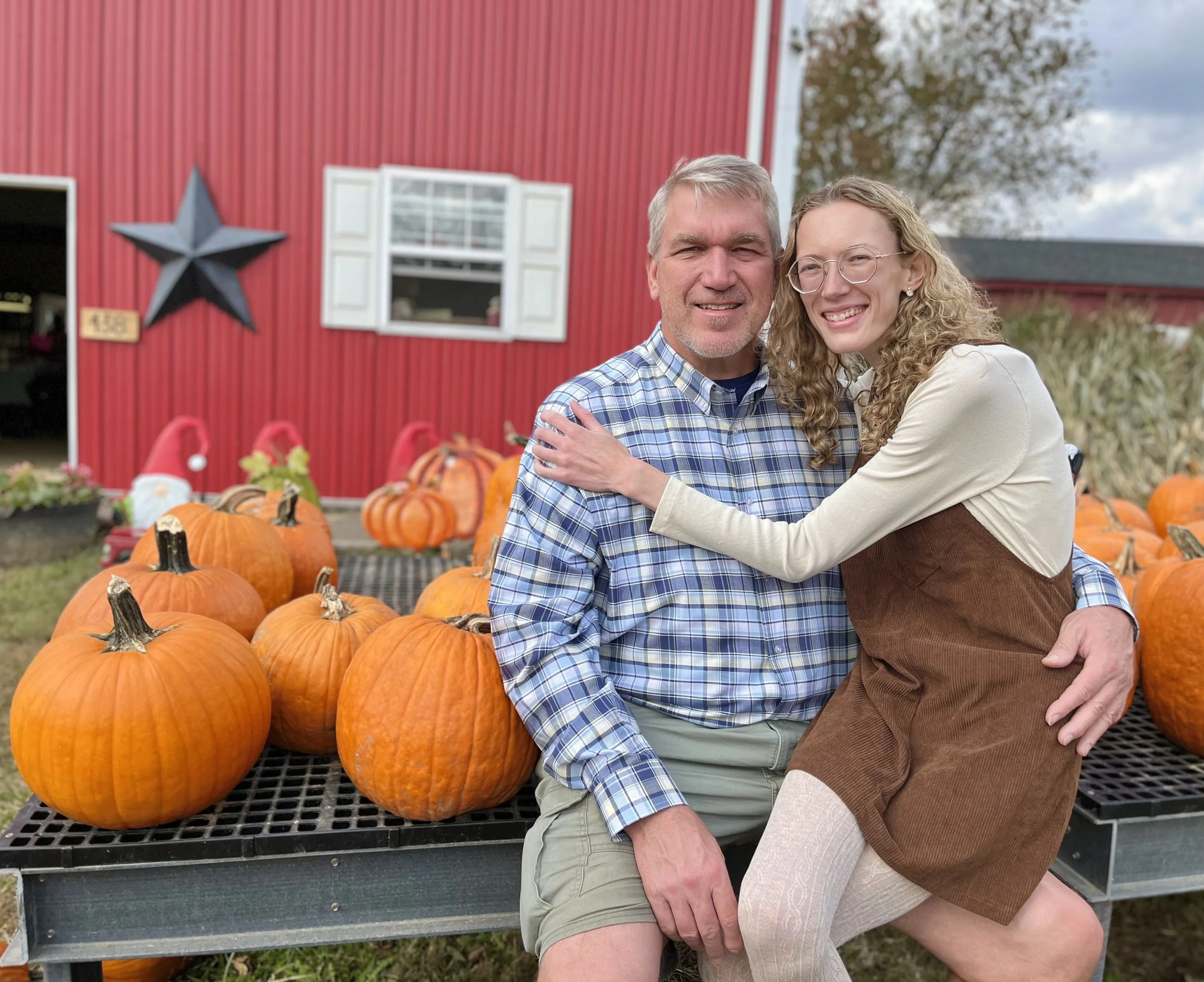 A couple sitting on a black metal table surrounded by pumpkins outdoors with a red barn and trees in the background.
