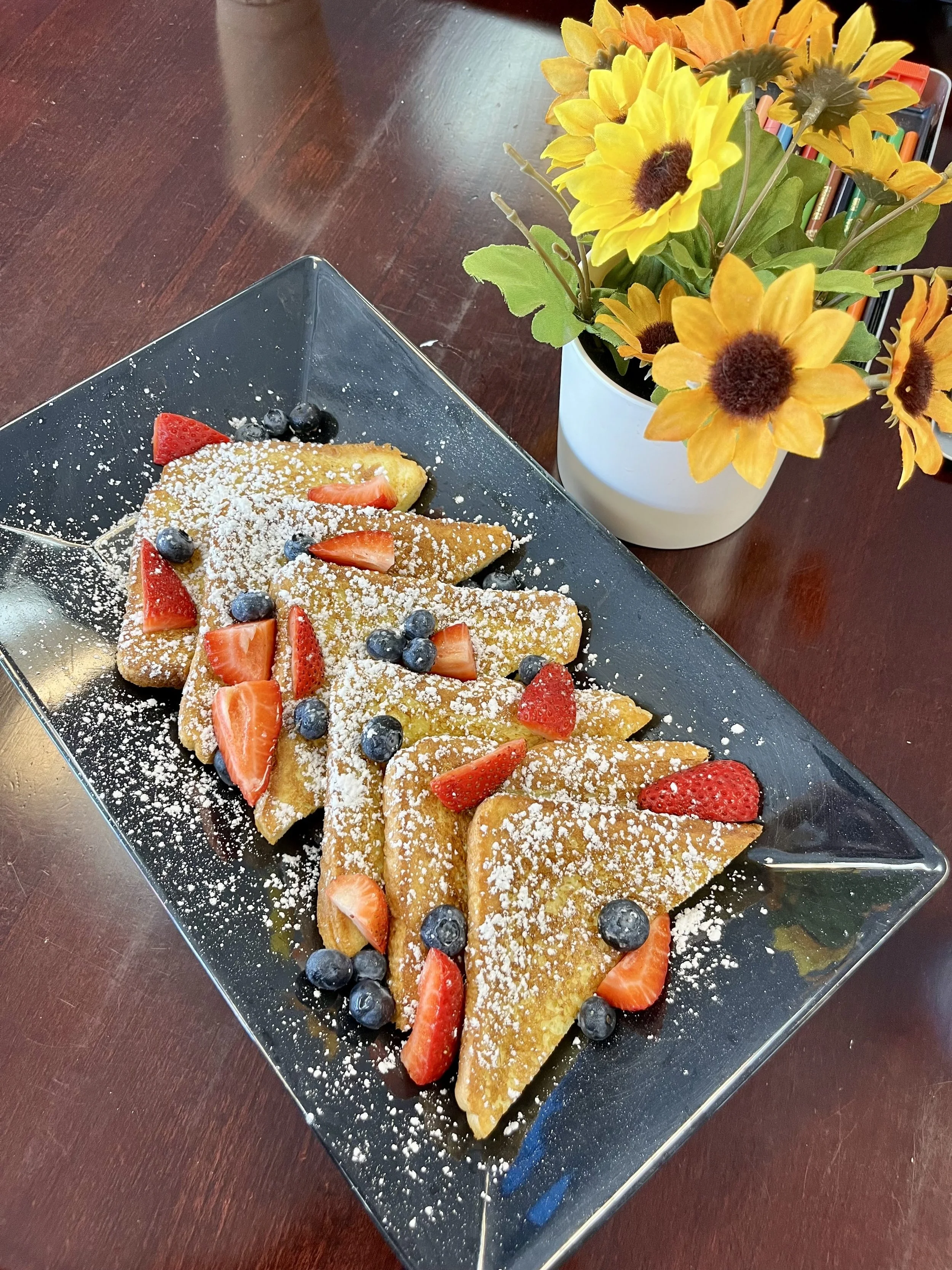 Plate of French toast topped with powdered sugar, sliced strawberries, blueberries, and garnished with a bouquet of yellow and orange sunflowers.
