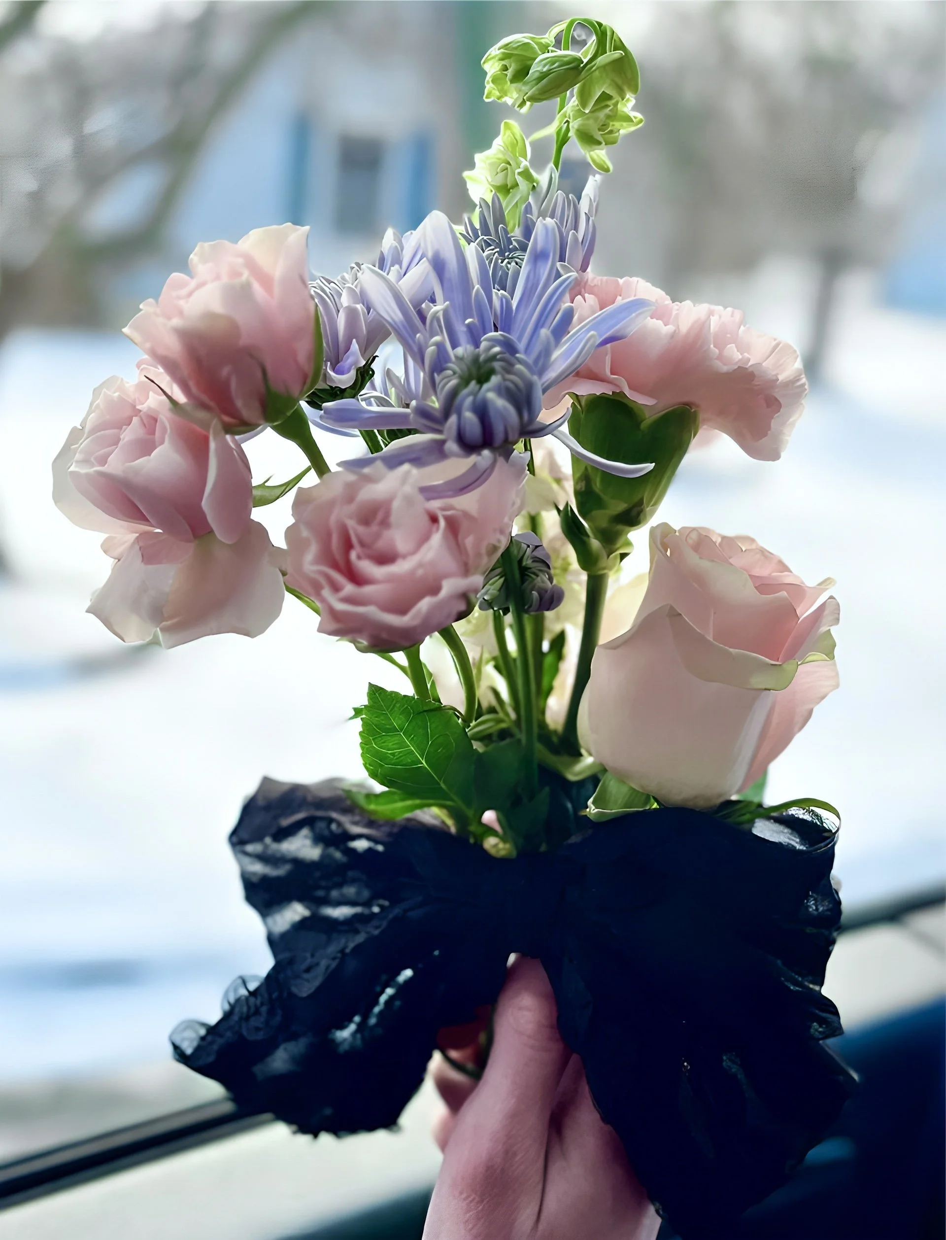 A hand wearing a black glove holding a small black pot with a bouquet of flowers indoors, with a blurred background of a window and trees.