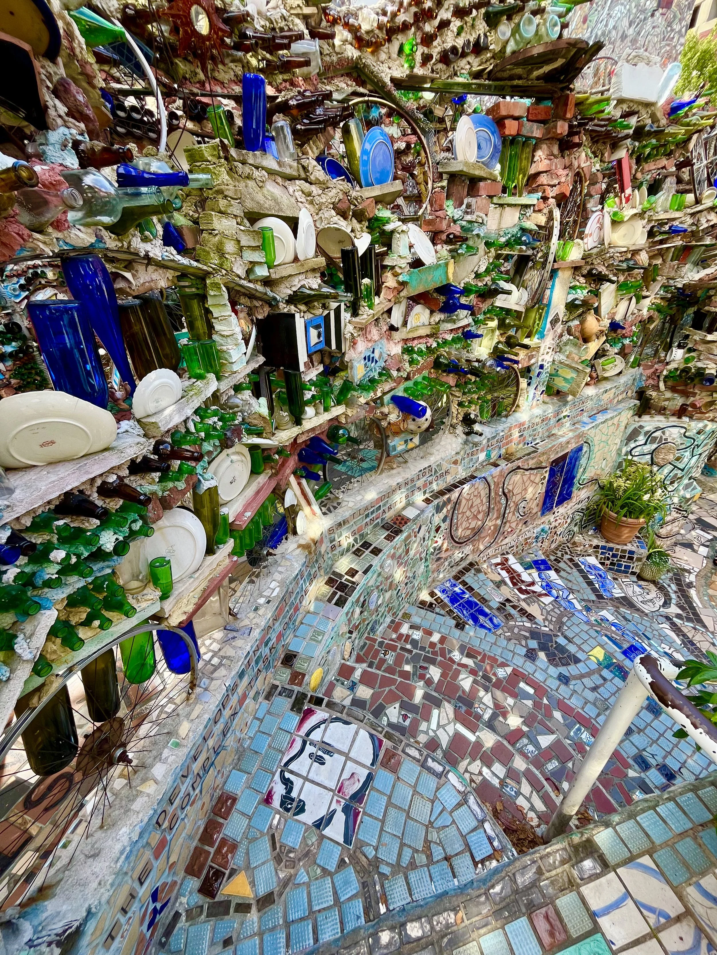 Colorful mosaic wall and floor decorated with various glass bottles, plates, and tiles, forming an artistic and vibrant outdoor installation.
