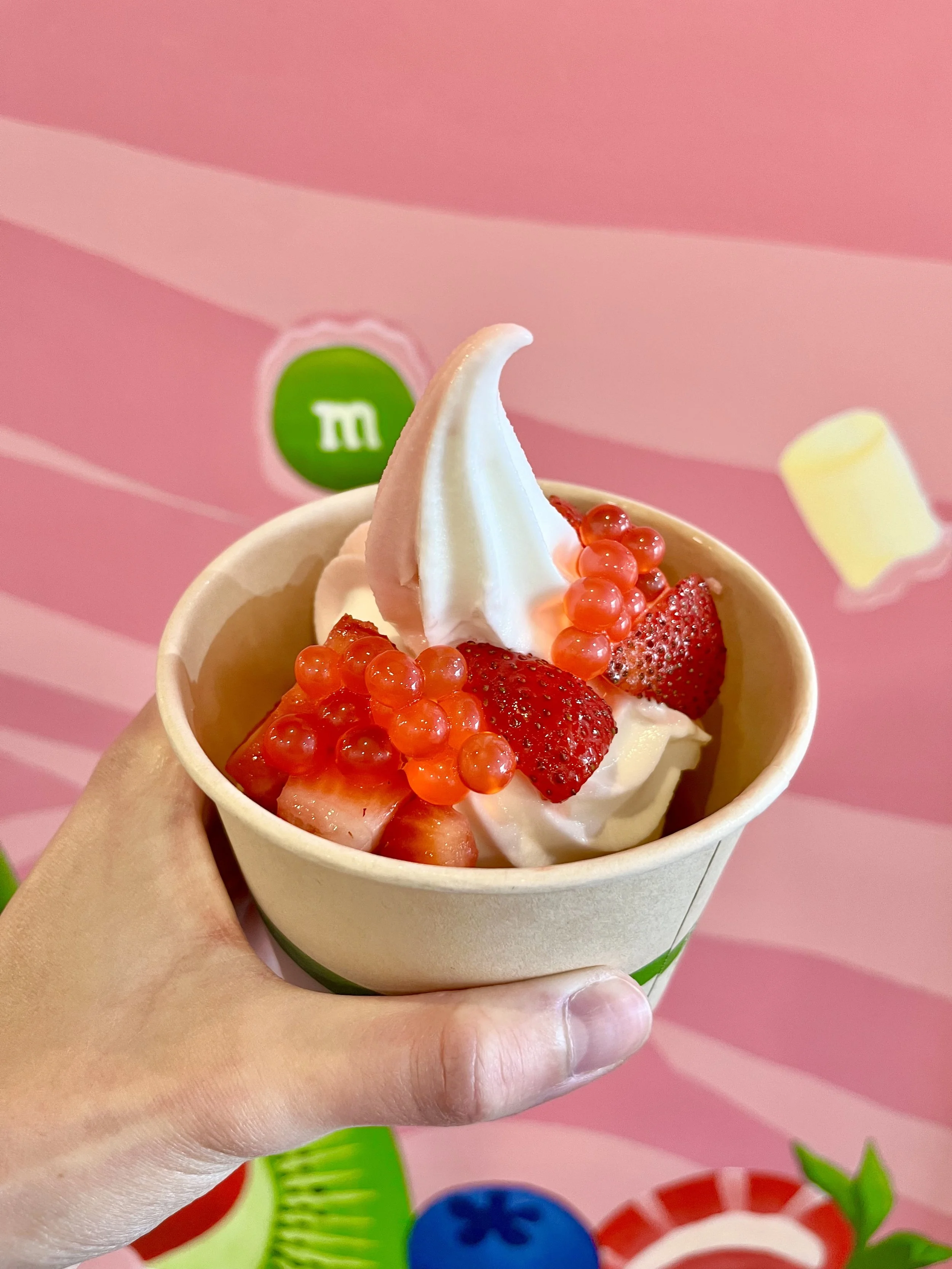 Hand holding a cup of frozen yogurt topped with strawberries, red currants, and slices of strawberry against a pink background with colorful fruit illustrations.
