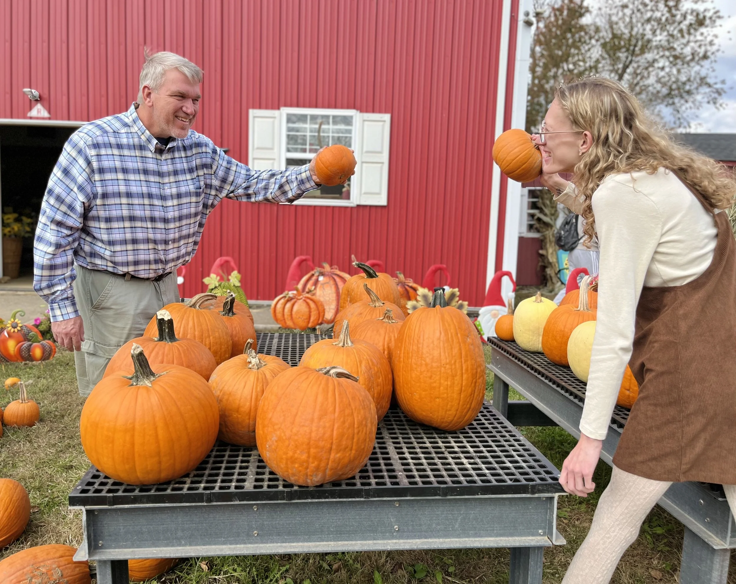 A man and a woman at a pumpkin stand, each holding a pumpkin near their noses, smiling. The background features a red barn and fall decorations.