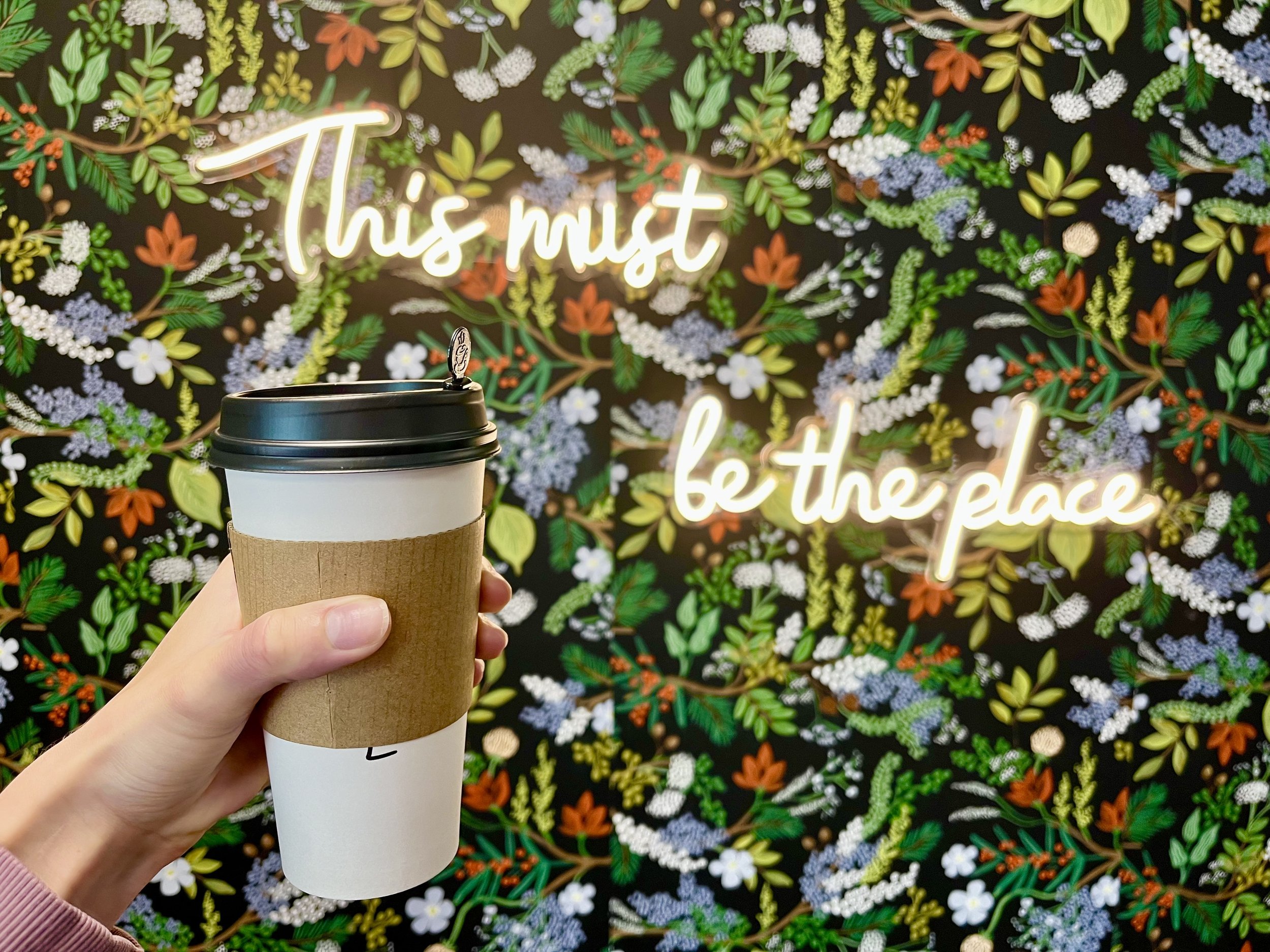 A hand holding a paper coffee cup with a black lid in front of a colorful floral and leafy background. The background features a neon sign reading 'This must be the place' amid various vibrant flowers and greenery.
