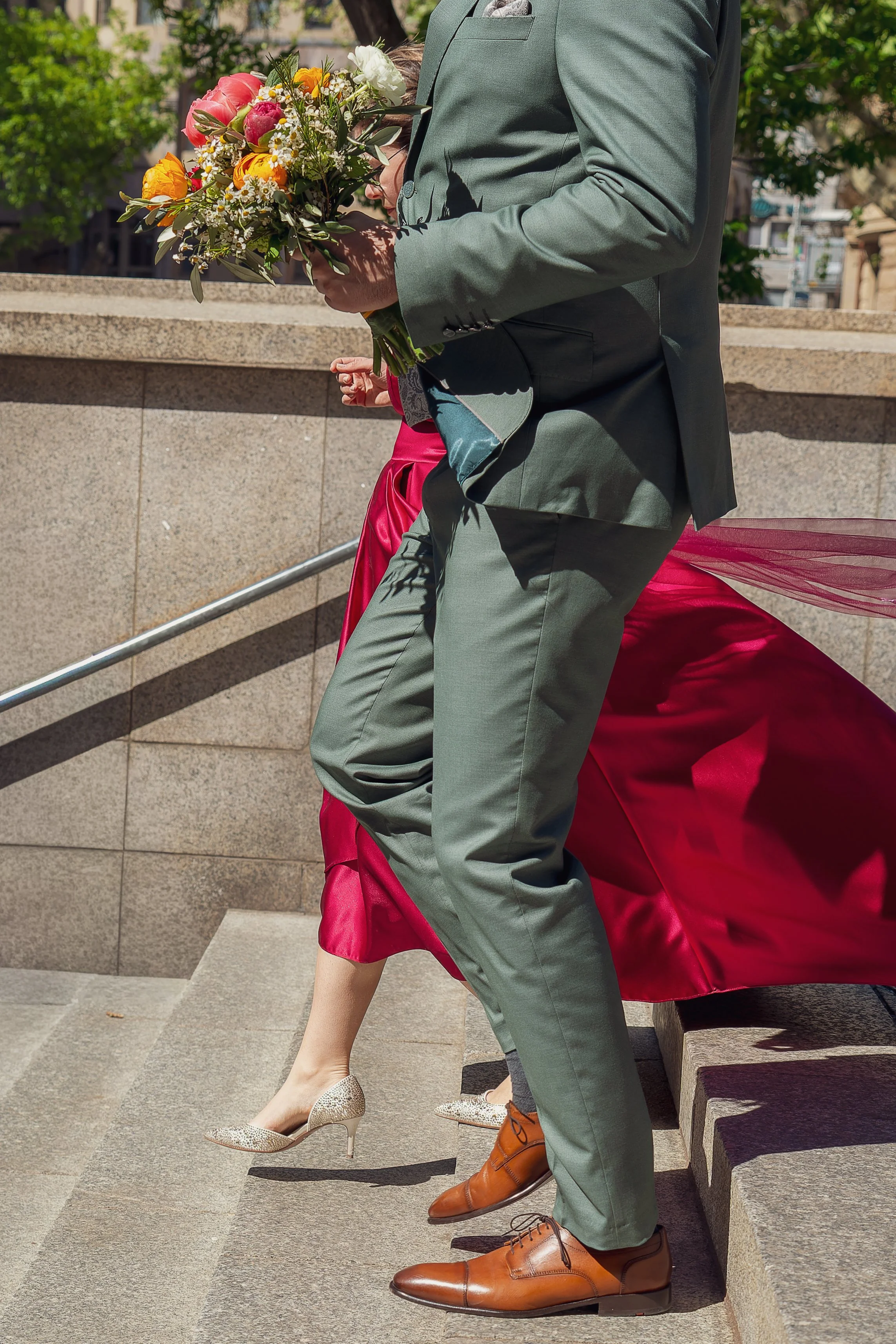 Menschen sitzen auf einer Bank im Freien, eine Person in einem grauen Anzug hält einen bunten Blumenstrauß, die Beine von mehreren Menschen sind sichtbar, sie tragen elegante Schuhe, und im Hintergrund sind Bäume und Gebäude zu sehen.