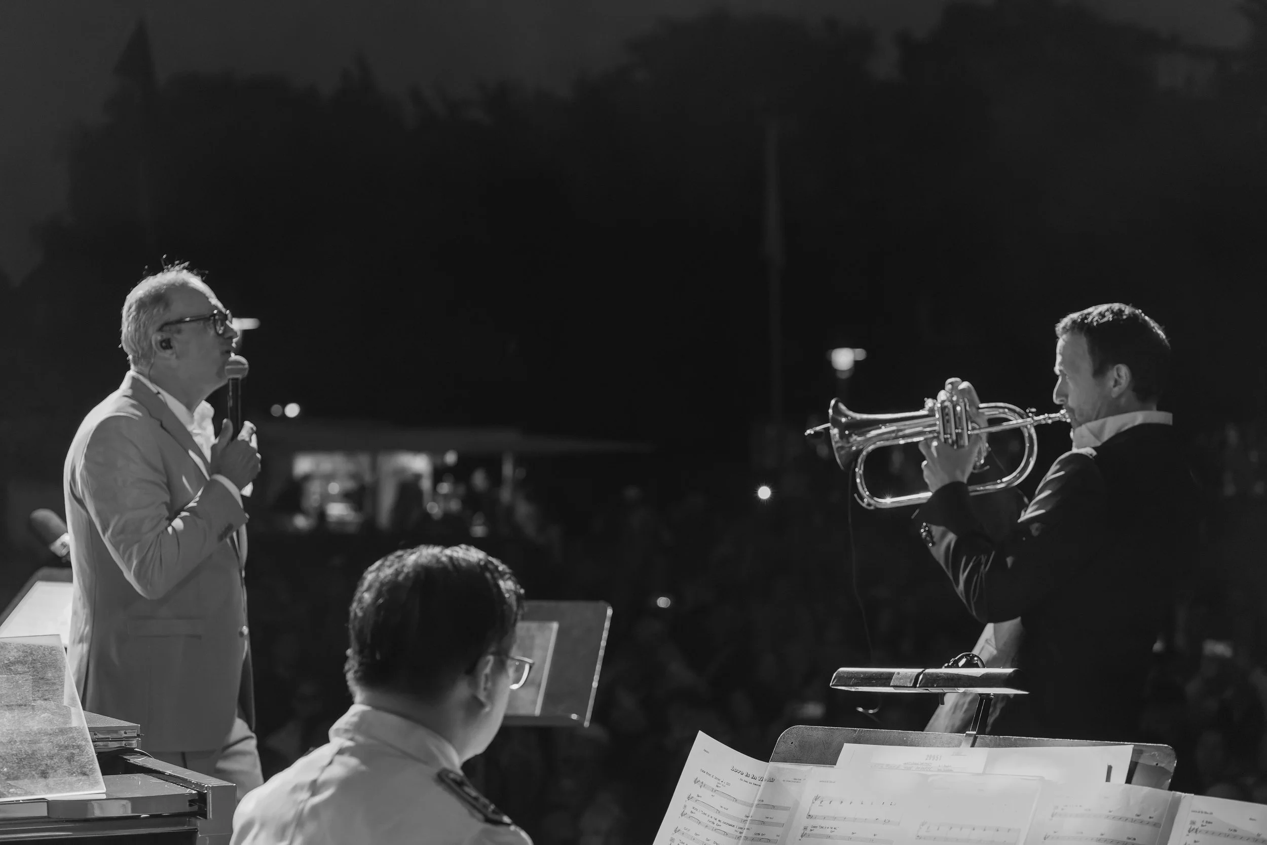Zwei Musiker während eines nächtlichen Konzerts im Freien, einer singt mit einem Mikrofon, der andere spielt Trompete.
