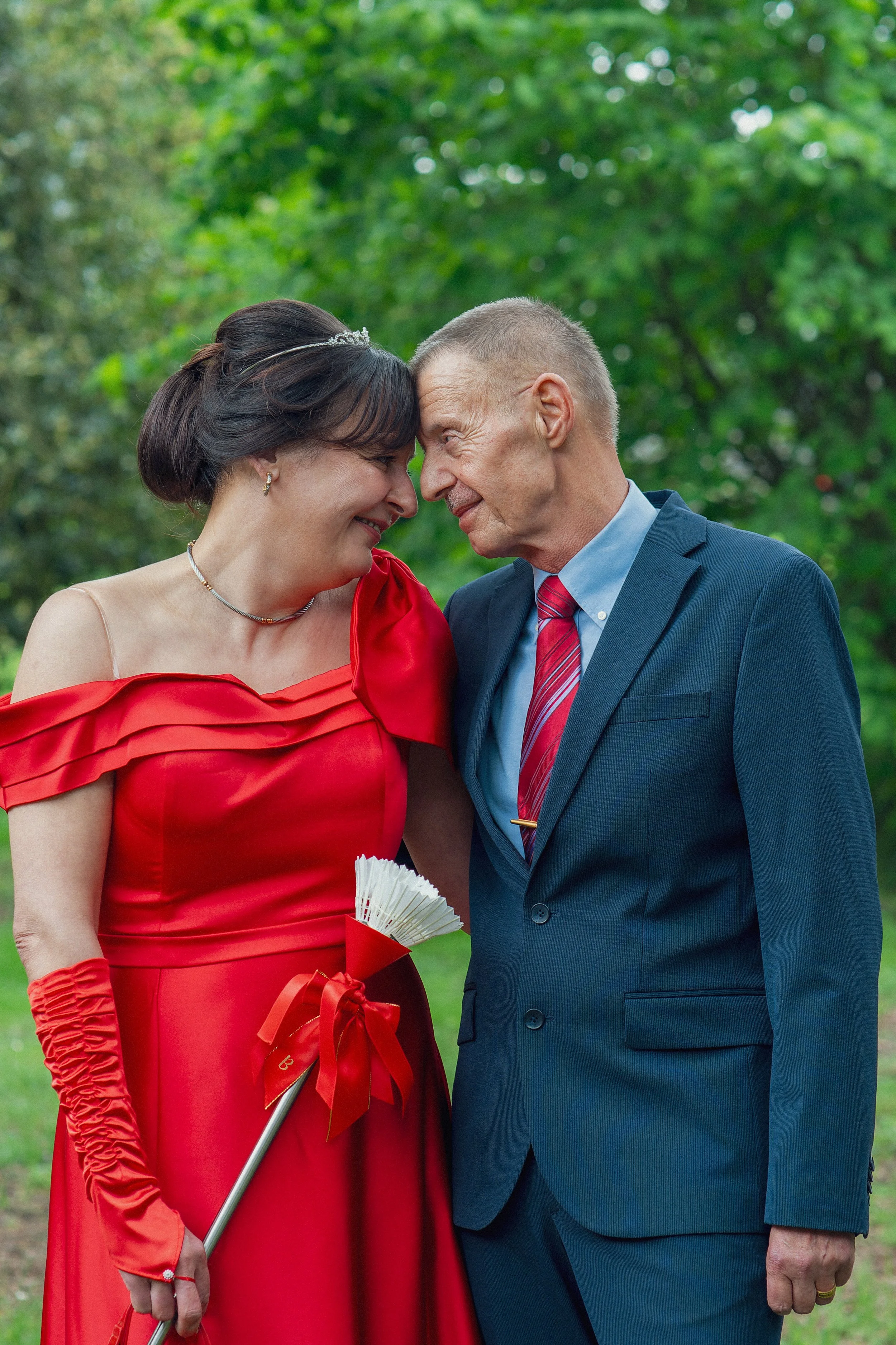 Ein Mann in einem dunklen Anzug und eine Frau in einem roten Kleid, die sich die Stirn berühren, im Freien auf einer grünen Wiese.