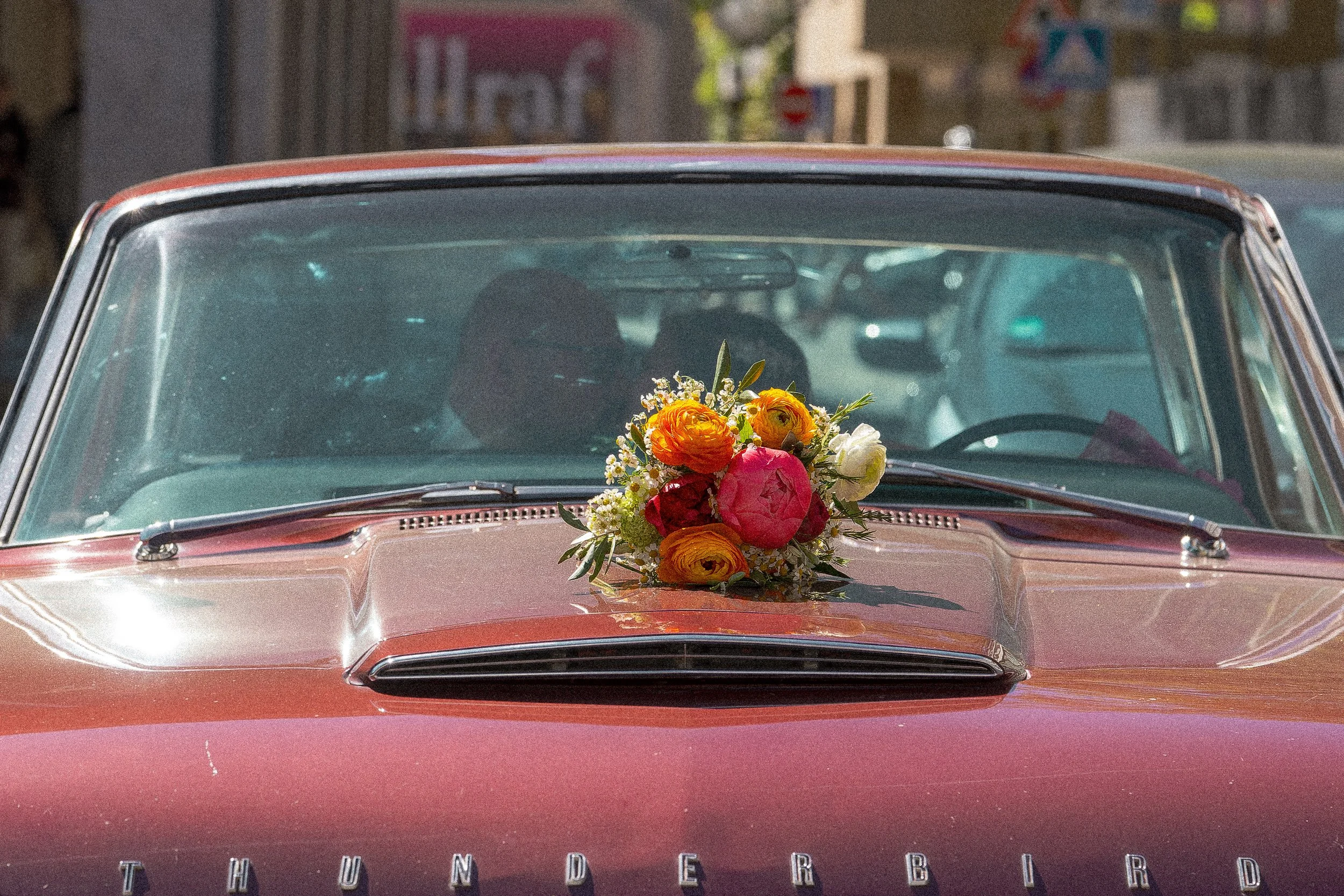 Ein rotes Auto mit einem Blumenstrauß auf der Motorhaube, aufgenommen bei Tageslicht auf einer Straße.