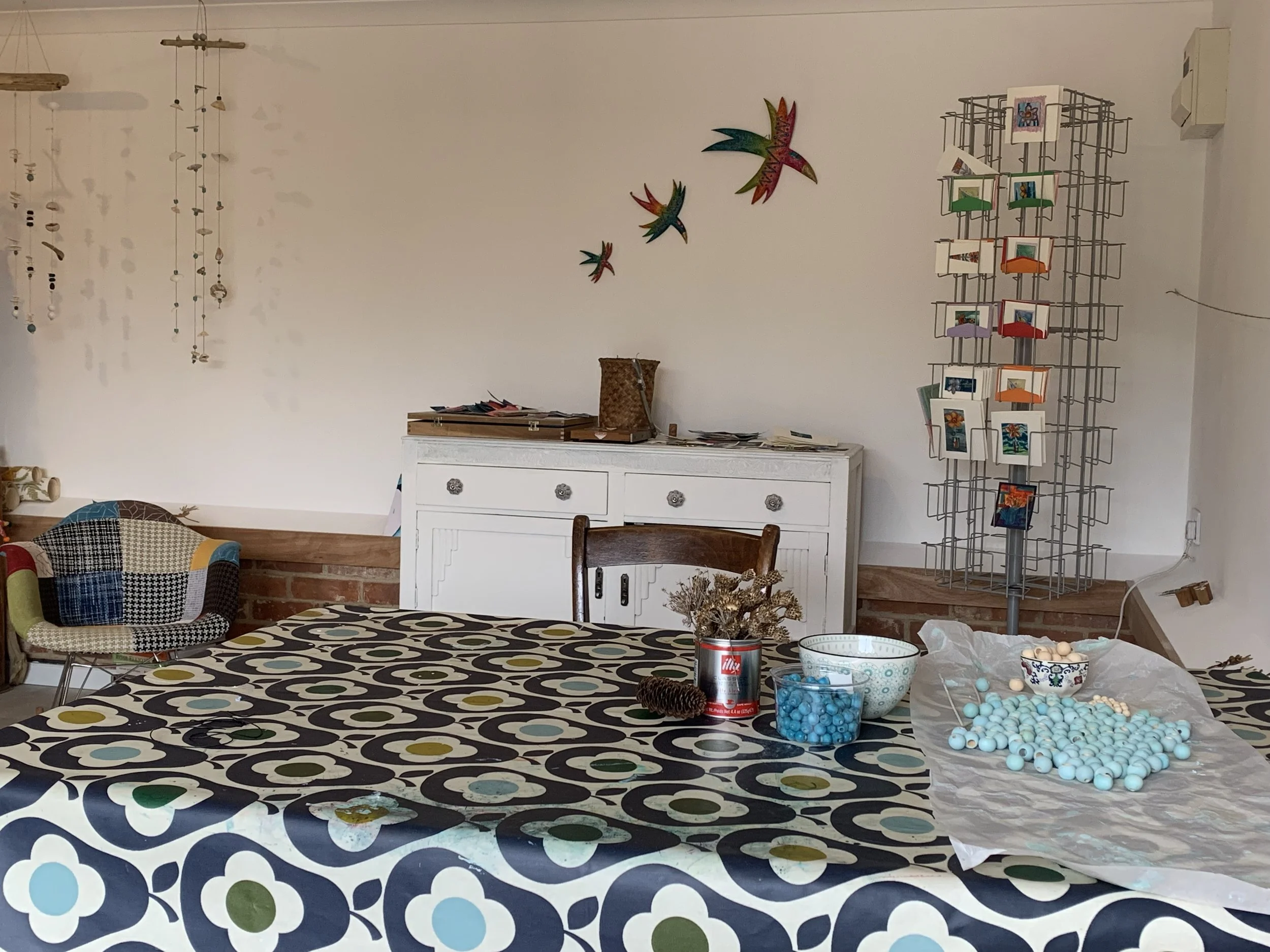 Interior of a room with a white dresser, colorful wall decorations of flying birds, a wire card display stand with greeting cards, and a table with various small blue balls and bowls, along with a can and some paper items.