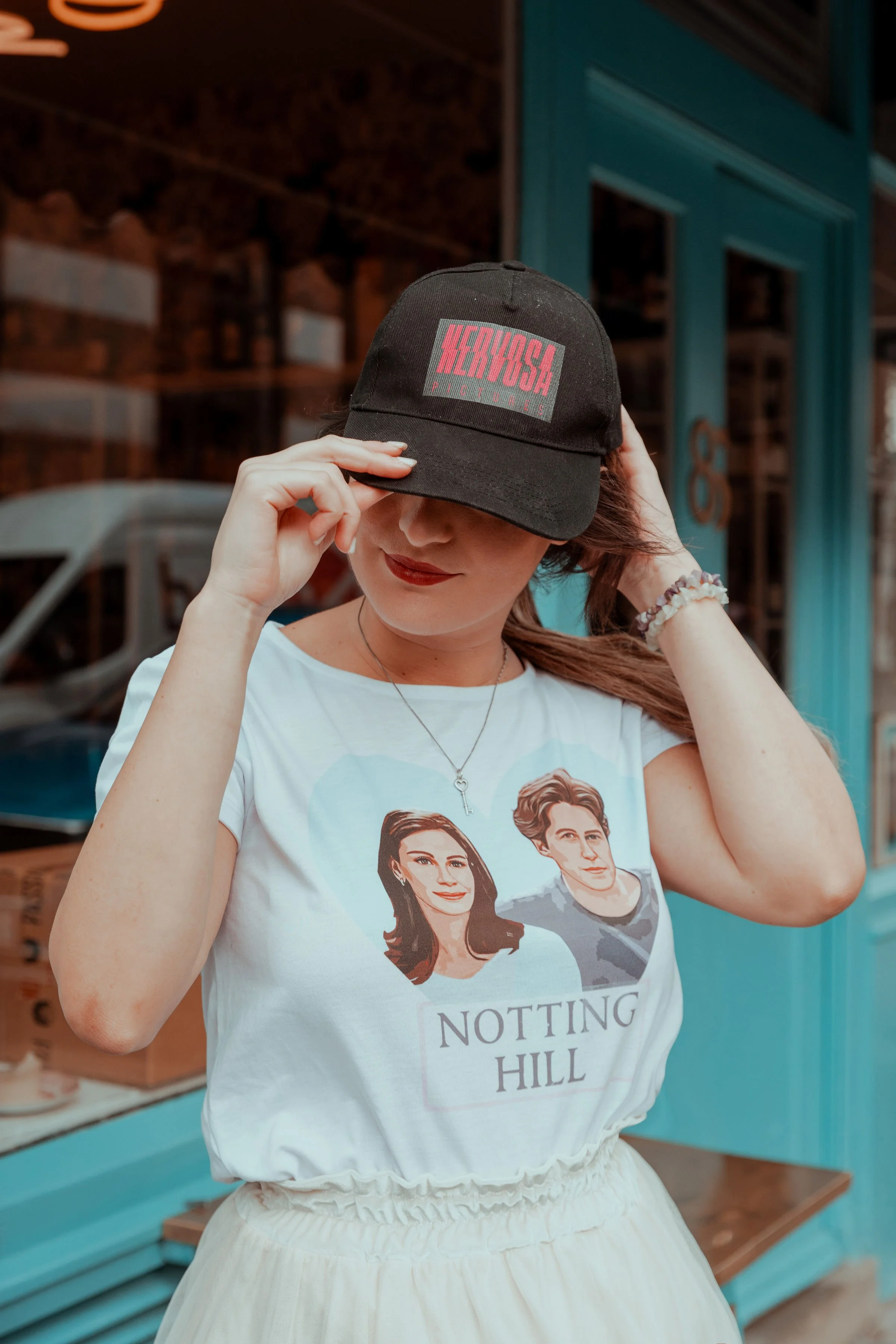 A woman wearing a black baseball cap with red and gray lettering, covering her face. She is also wearing a white T-shirt with illustrated portraits of a woman and a man, and the text 'NOTTING HILL'.