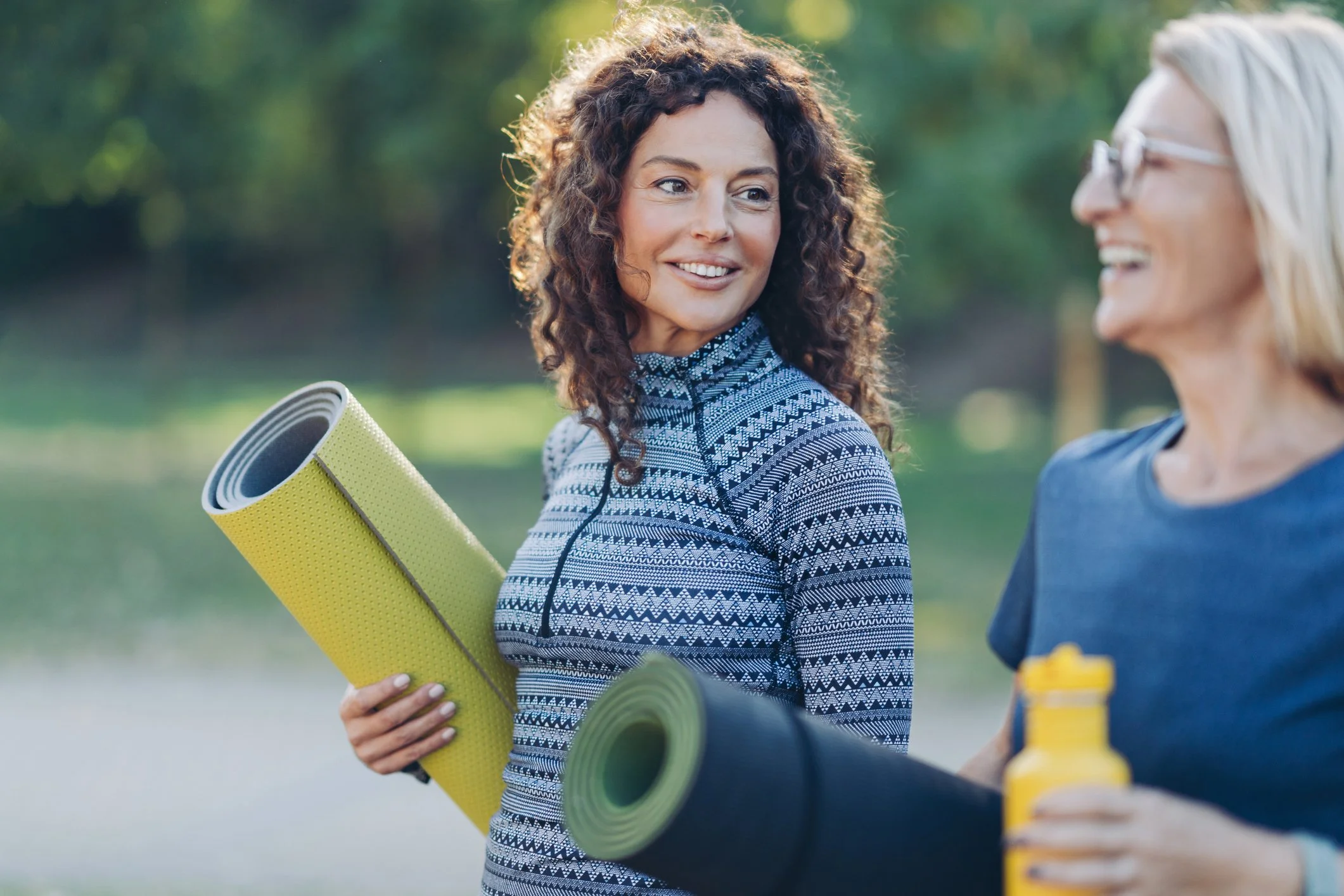 Two women are outdoors, smiling and chatting. One woman has curly hair, is wearing glasses, a patterned long-sleeve shirt, and is holding a rolled-up yoga mat. The other woman, partially visible, has straight blonde hair and is wearing a blue shirt. There are yellow and black objects on a table or surface in the foreground, and a blurred background of green trees and sunlight.