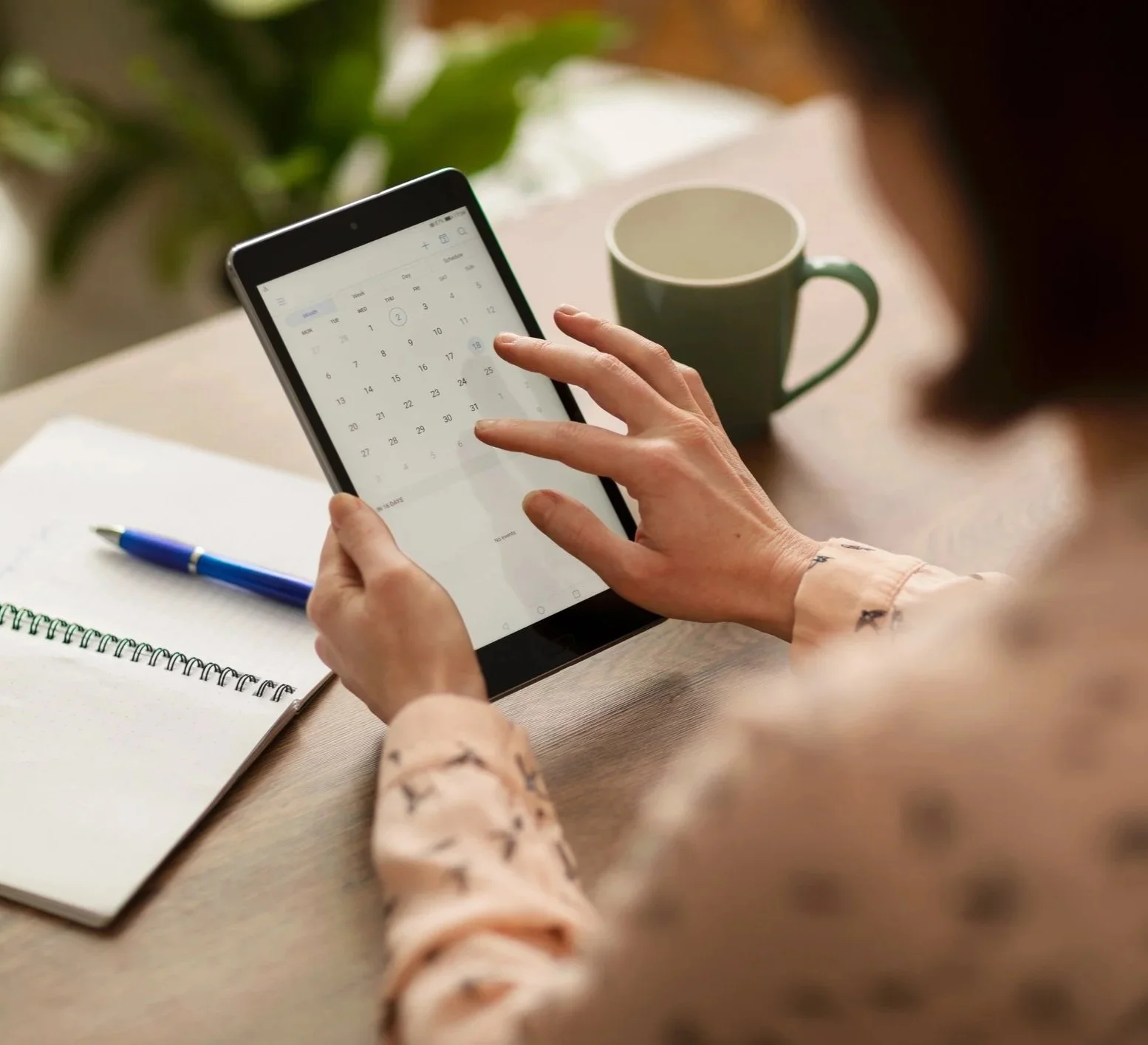 A person is holding a tablet displaying a calendar app, with a green coffee mug on the table next to them, alongside an open notebook and a blue pen.