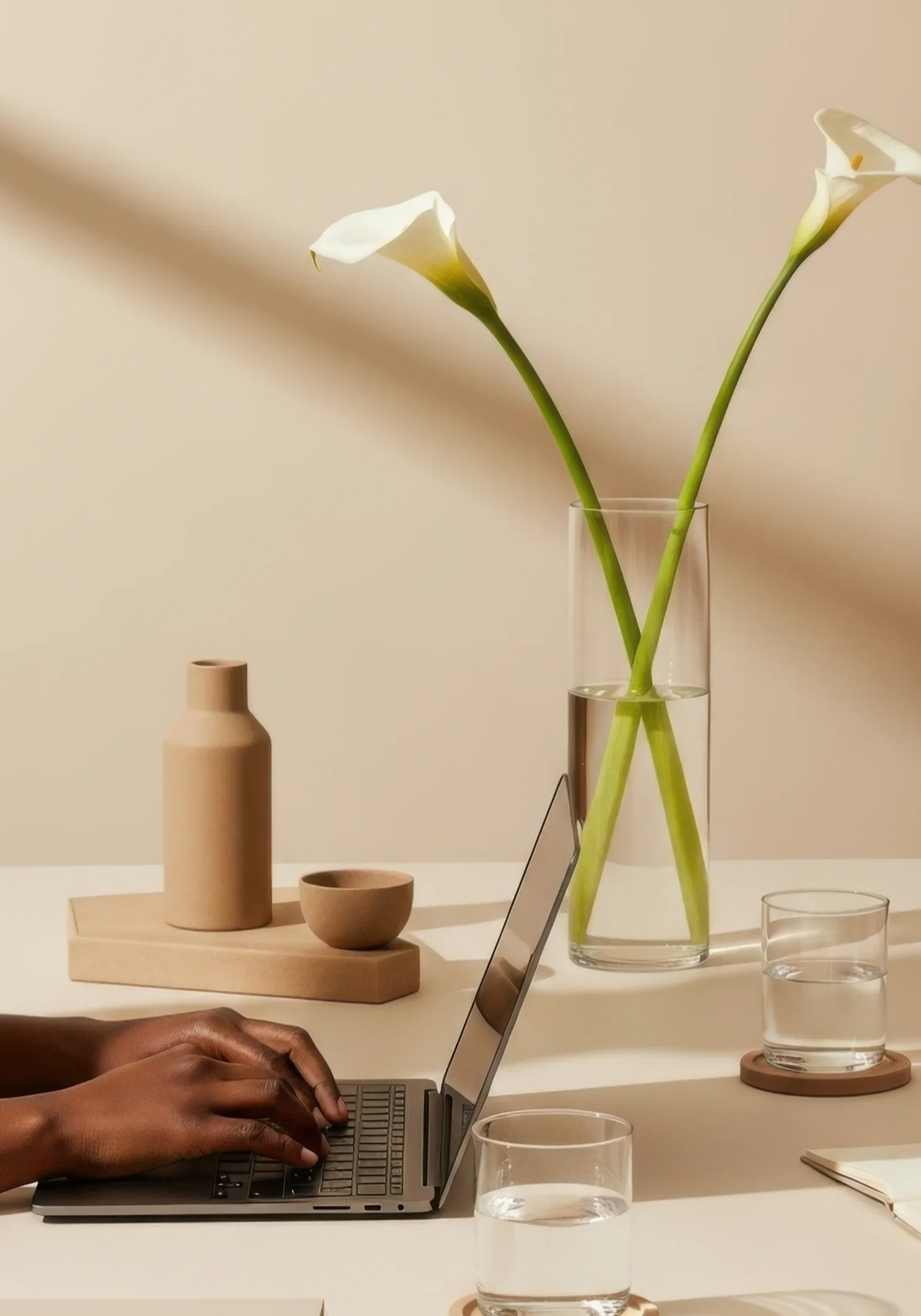 A person’s hands typing on a laptop keyboard at a white table. On the table are two glasses of water, one in front and one to the right. Behind the laptop are decorative objects, including a beige vase, a bowl, and a tall glass vase with white calla lilies.