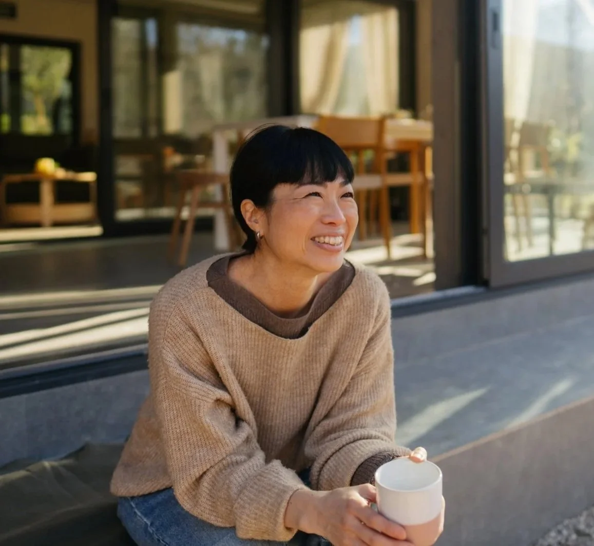 A smiling woman with short black hair sitting outside near a glass door, holding a white mug, dressed in a tan sweater and blue jeans, with a cozy and bright setting in the background.