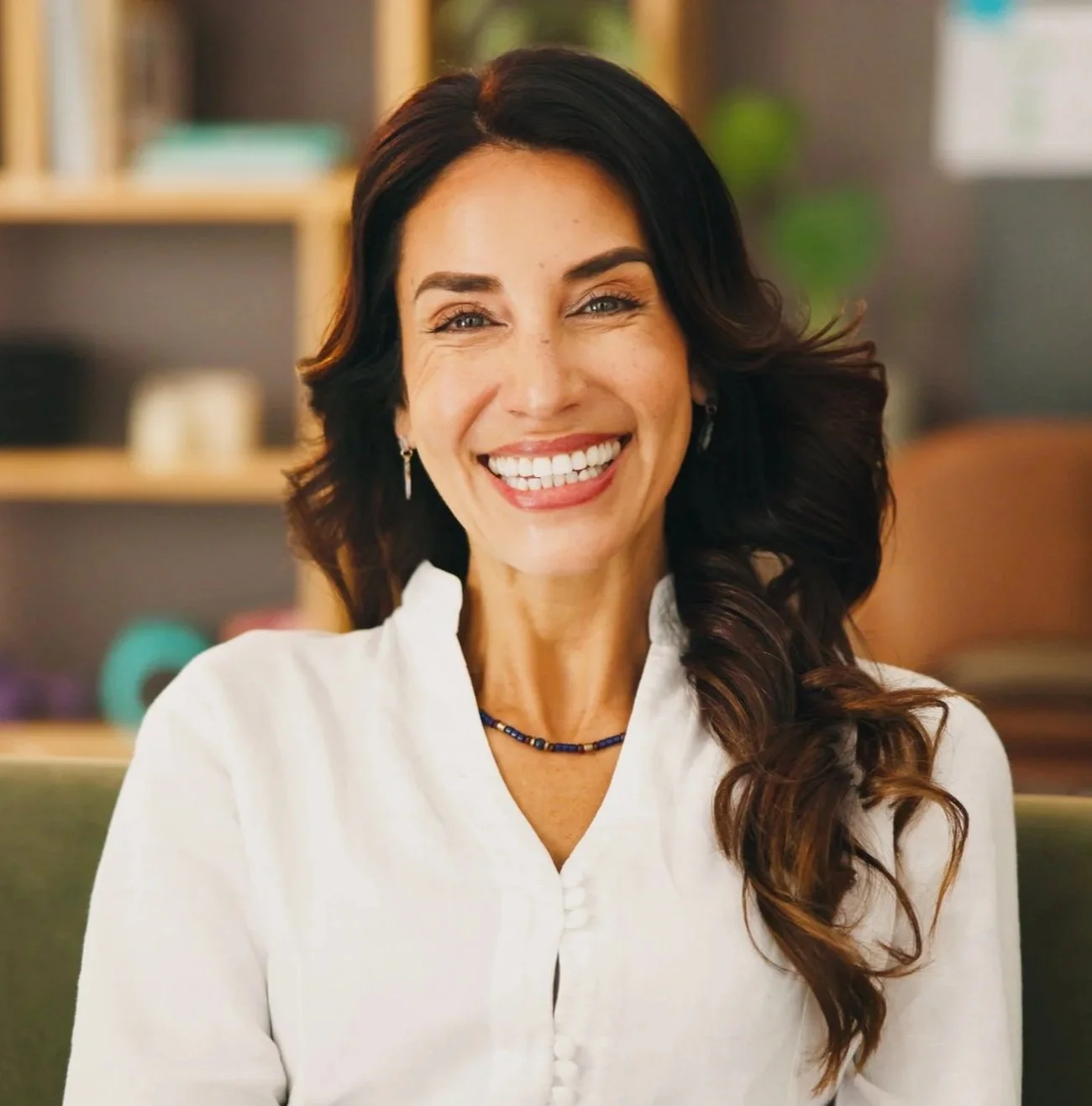 A woman with long dark hair and light skin, smiling broadly, wearing a white shirt, earrings, and a necklace, in a cozy, modern indoor setting.