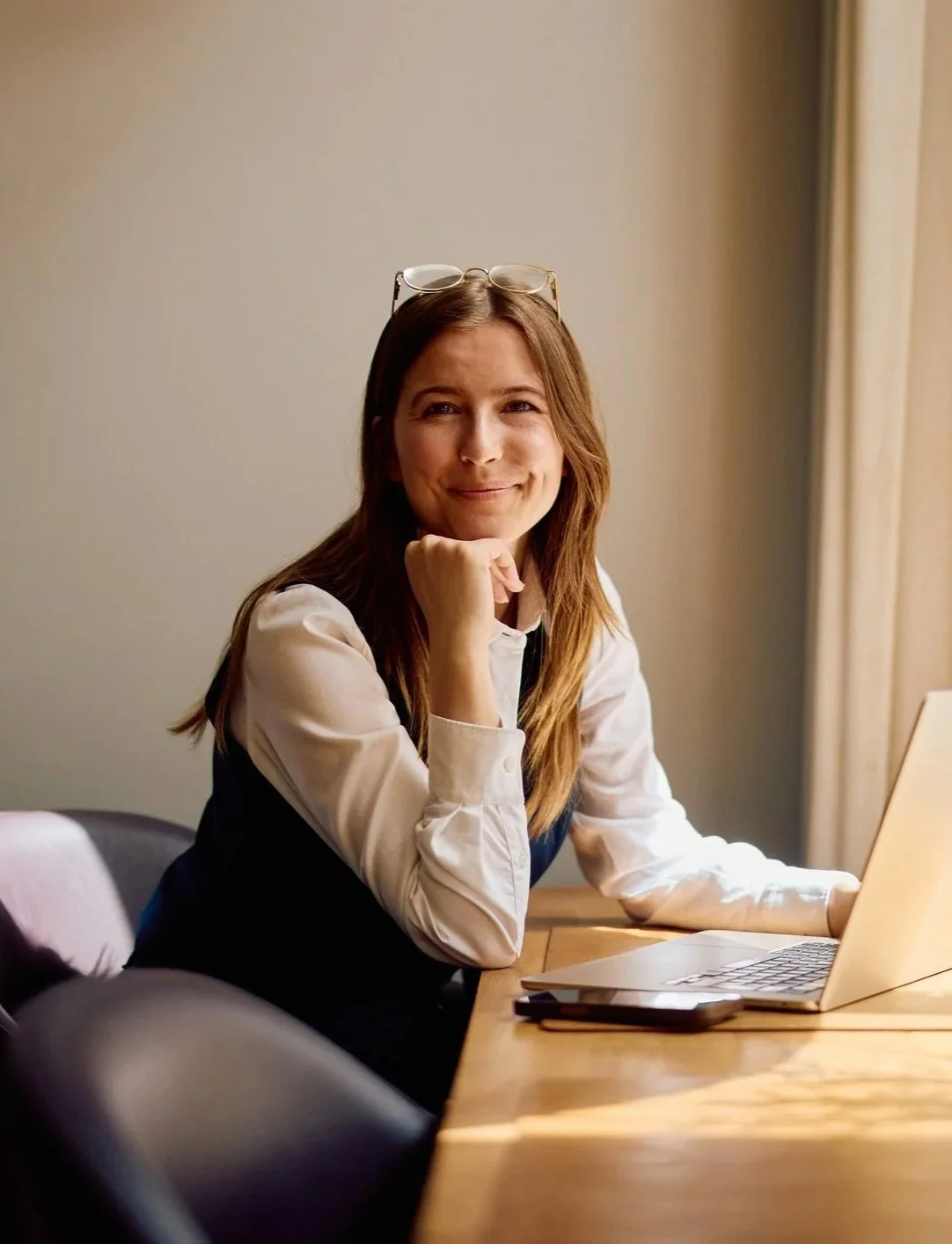 A woman with glasses on her head sitting at a wooden desk with a laptop, smiling at the camera.