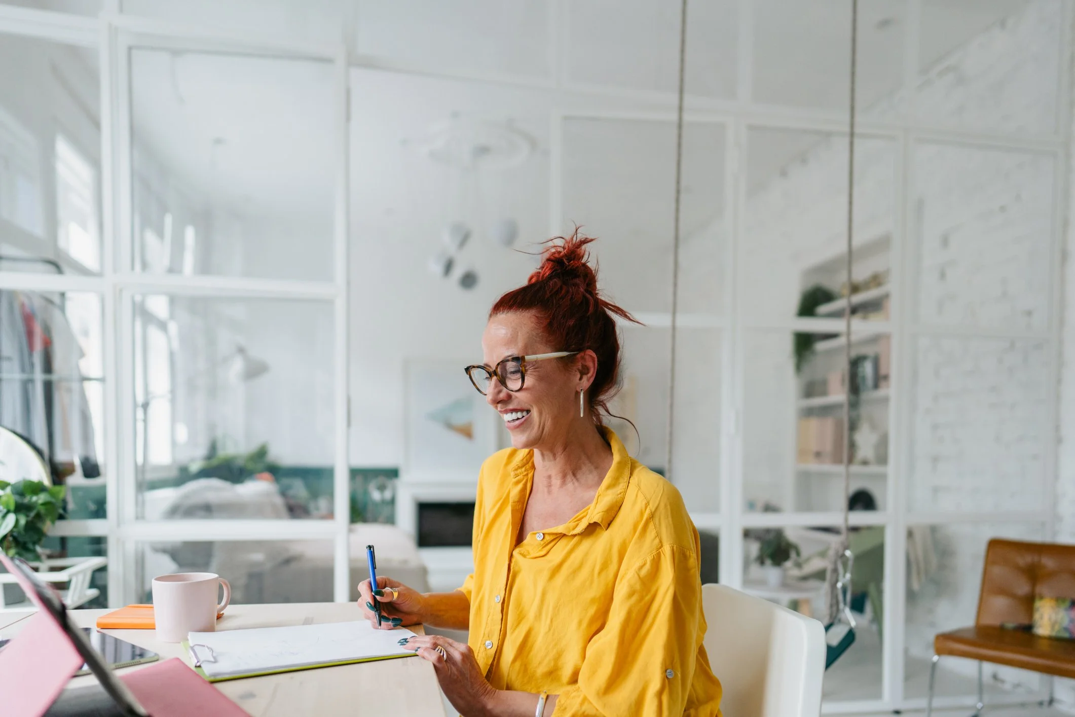 A woman with red hair in a bun, wearing glasses and a yellow shirt, smiling while sitting at a table in a bright room with white walls and windows.