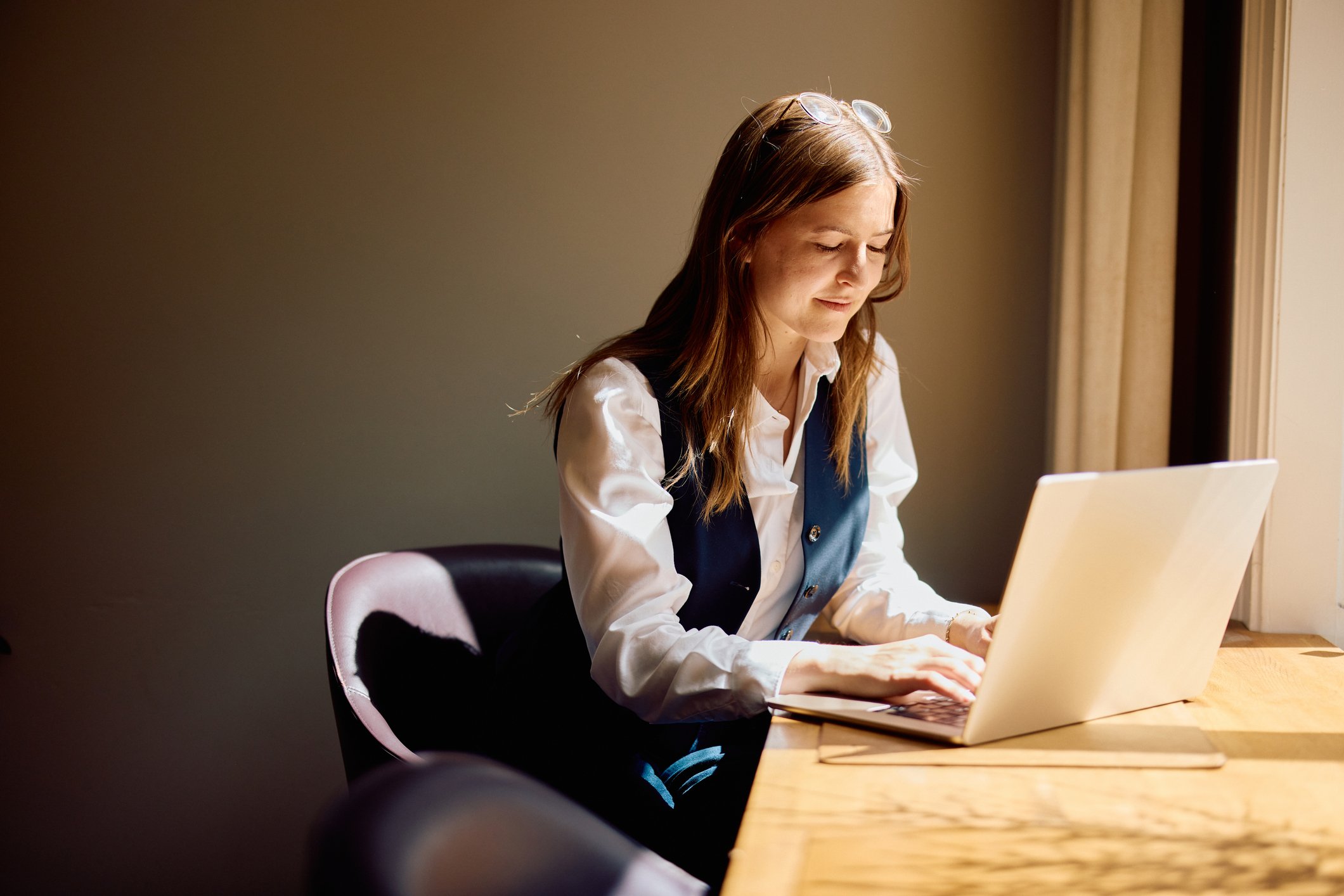 A young woman with long red hair working on a laptop in a well-lit room, sitting at a wooden desk.